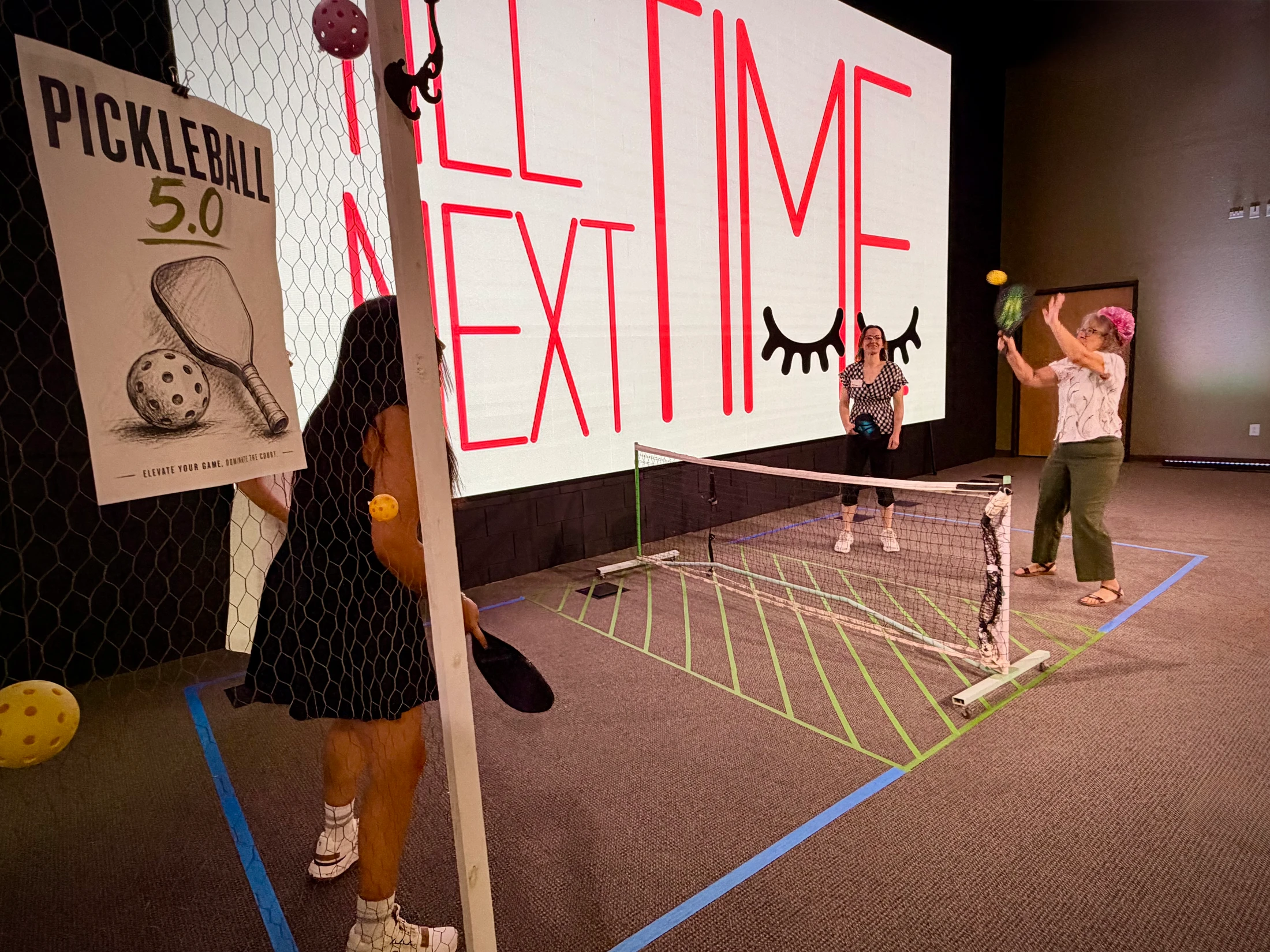 Three women play a game of pickleball on a makeshift indoor court made of tape, with a large screen in the background and a pickleball poster on a fence.