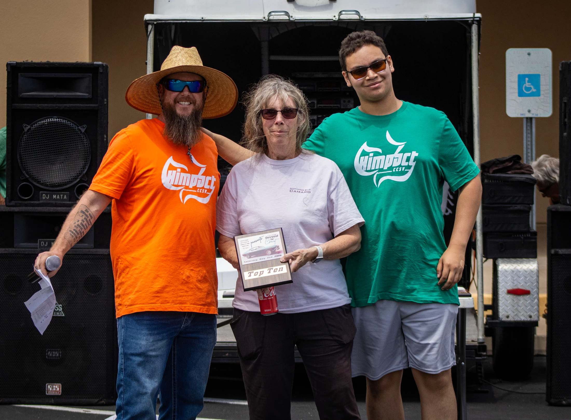 A woman holds a 'Top Ten' award plaque while posing for a photo between two men in matching t-shirts in front of large speakers.