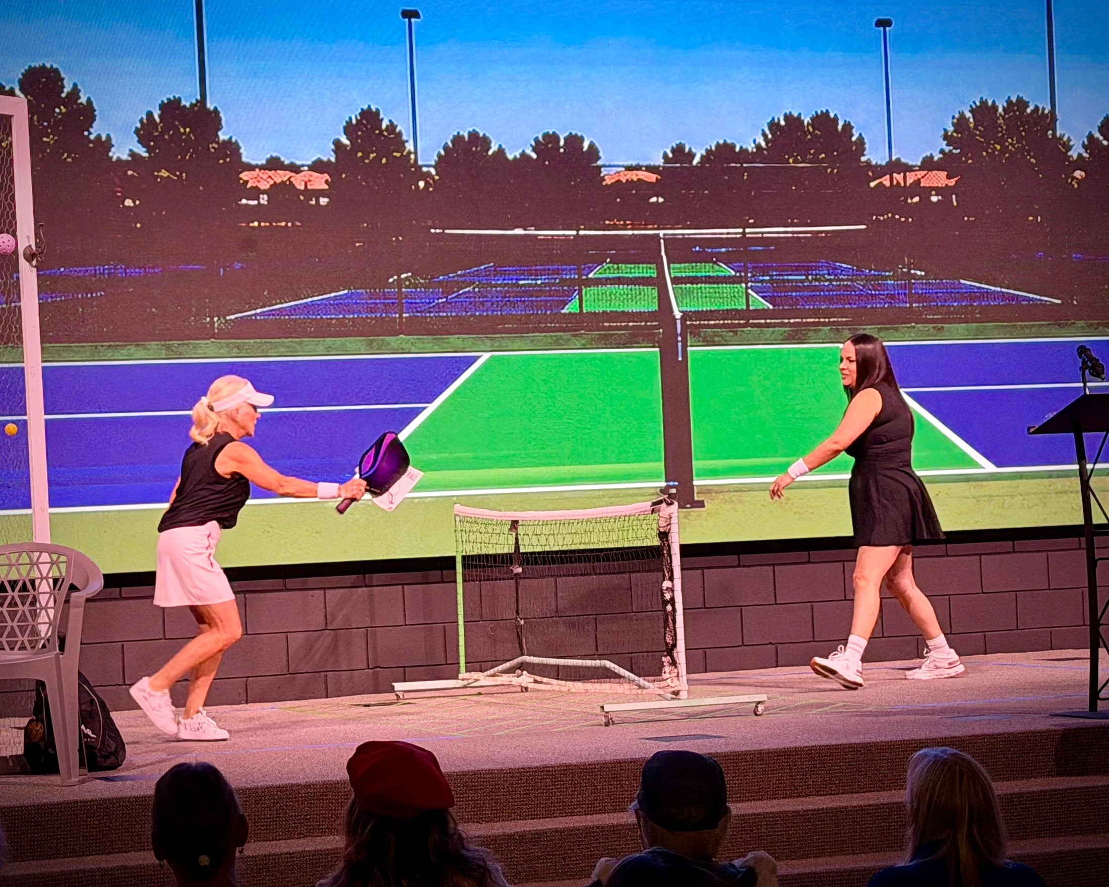 Two women play pickleball on a stage in front of a large screen showing a court. An audience watches from below as one woman hits the ball with a paddle.