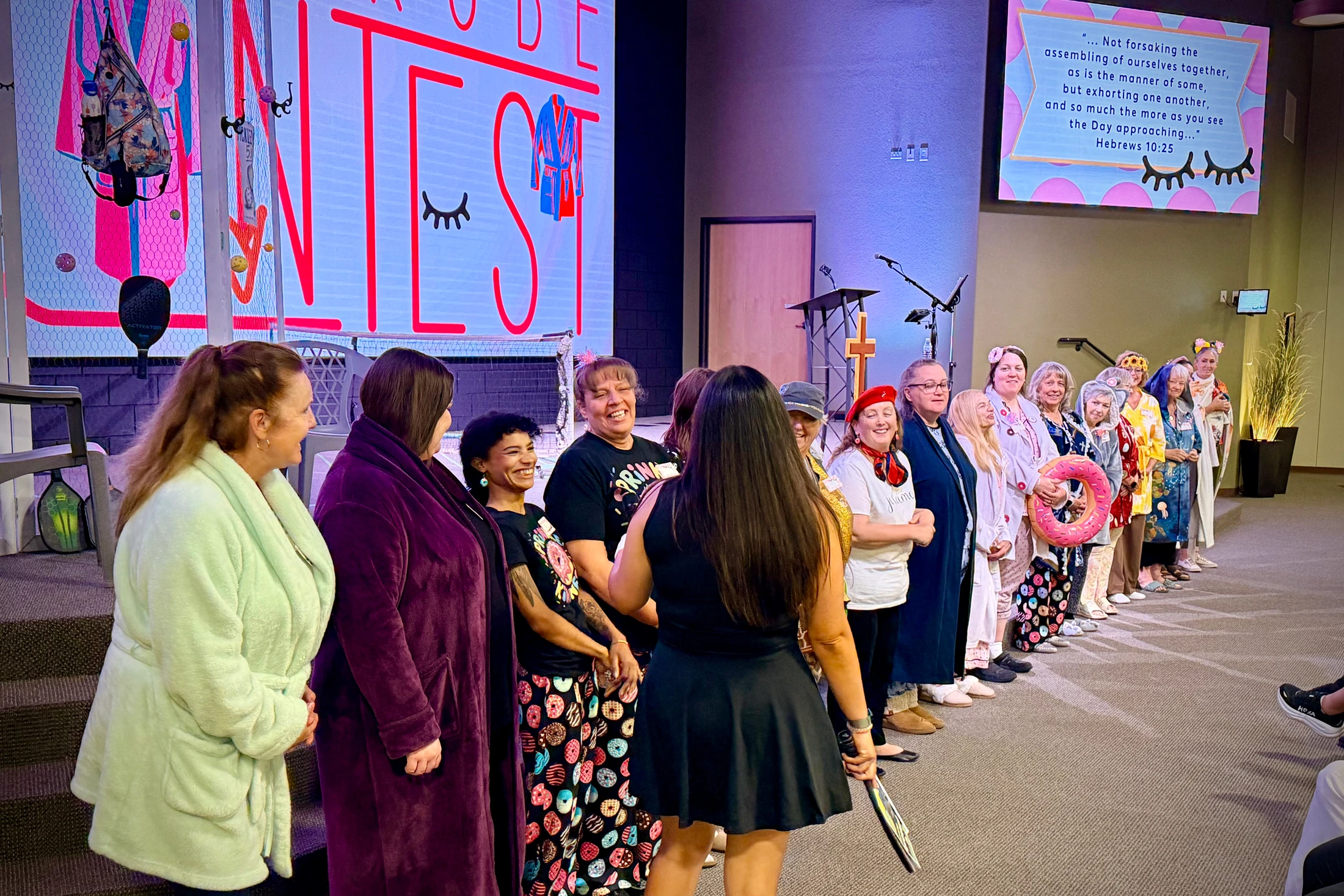 A long line of diverse women in colorful pajamas and robes stand smiling and laughing together inside a church auditorium during a fun-themed event.