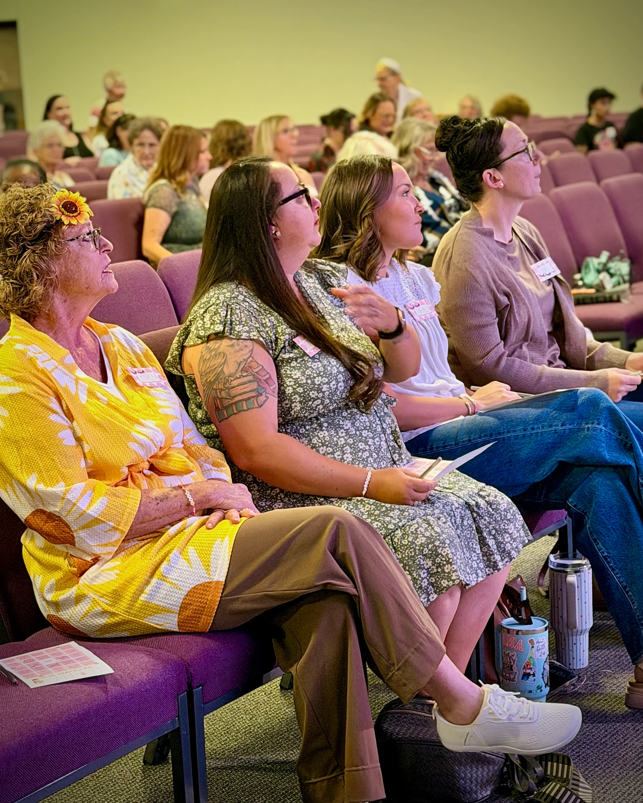 A group of women of different ages sit in purple chairs in an auditorium, listening attentively. One woman wears a bright yellow sunflower-themed outfit.