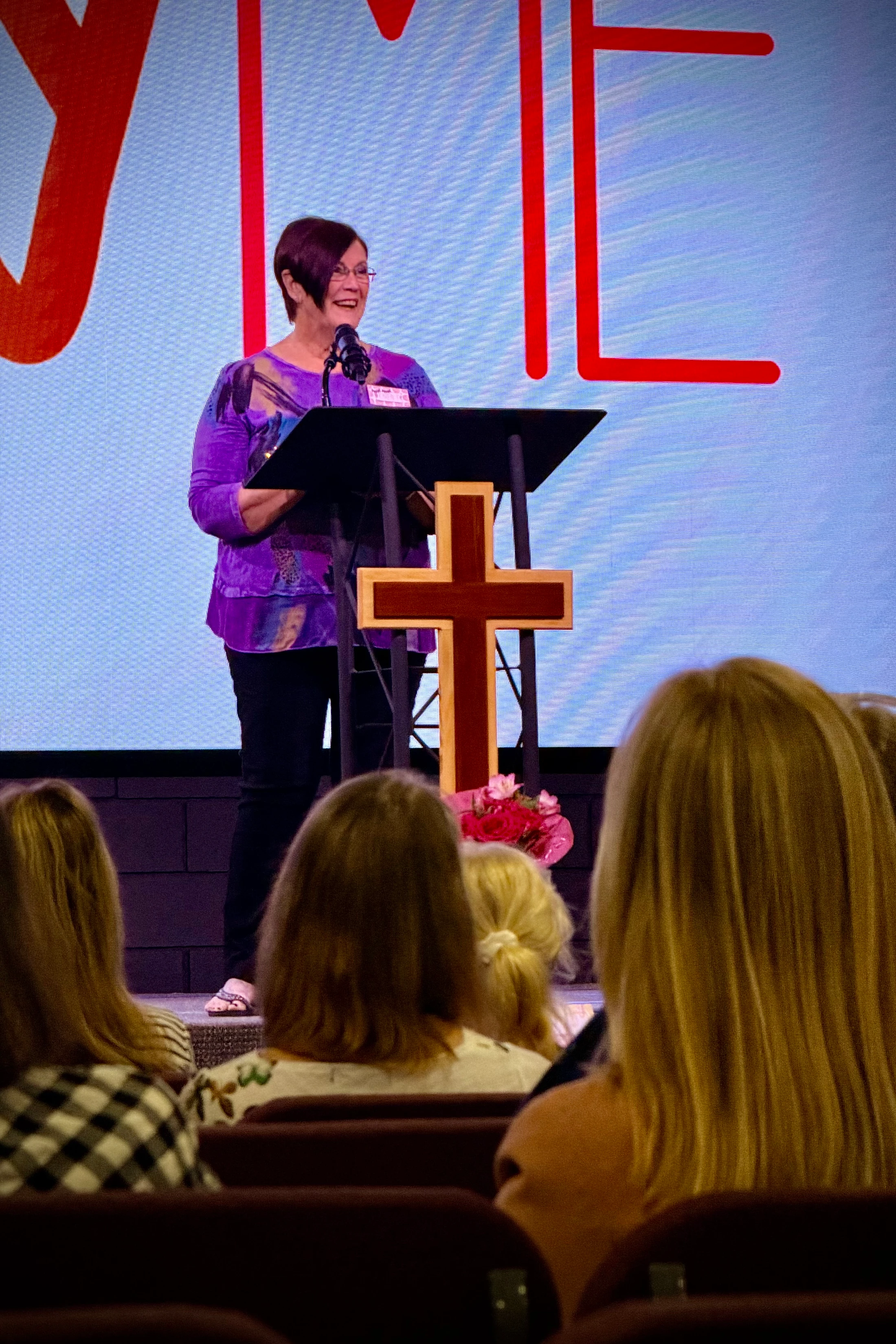 A smiling woman with glasses and a purple shirt speaks from a podium with a cross on it to an audience at a church event.