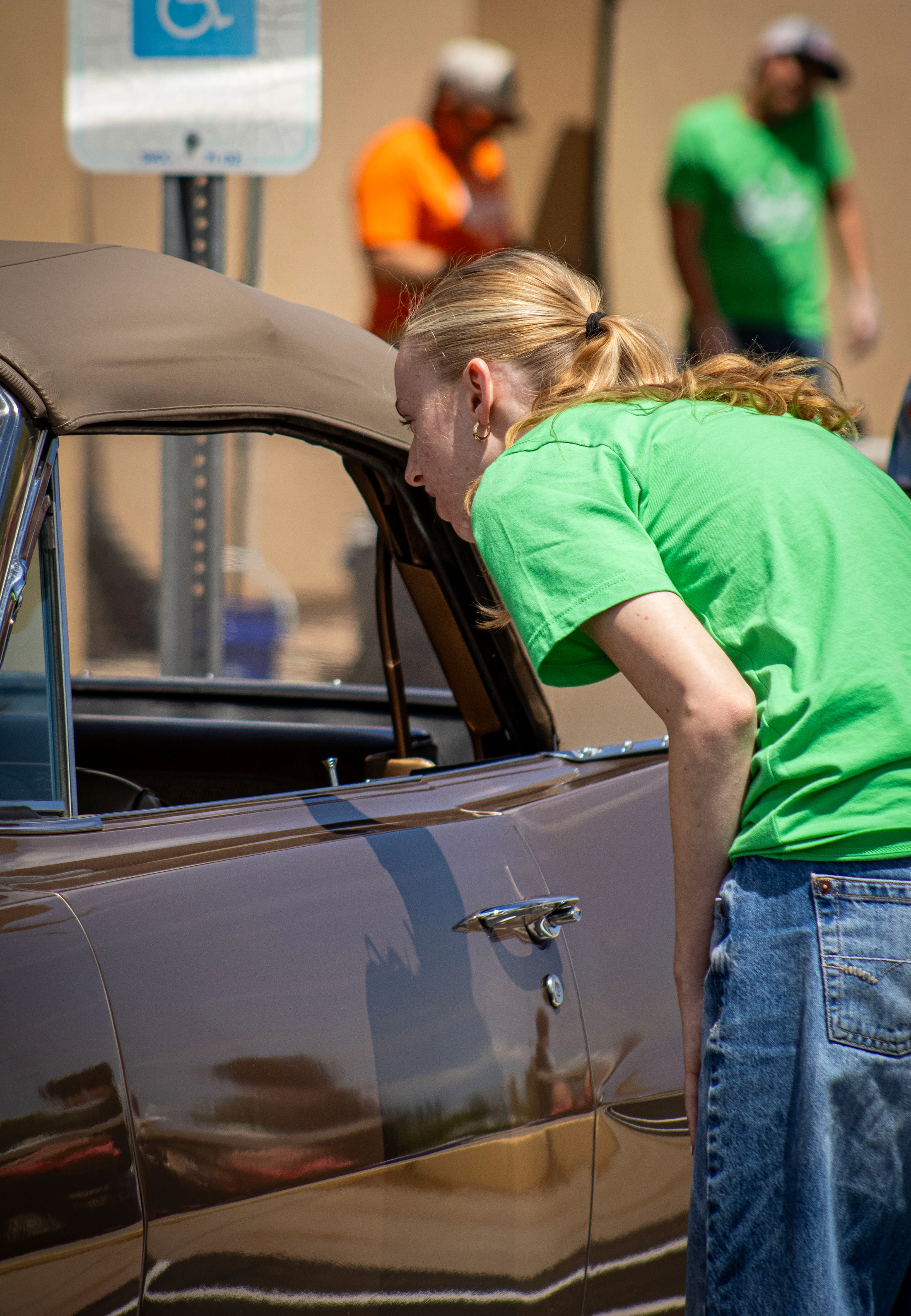 A young woman with blonde hair in a green shirt and jeans leans down to look through the open window of a shiny, dark brown classic convertible car.