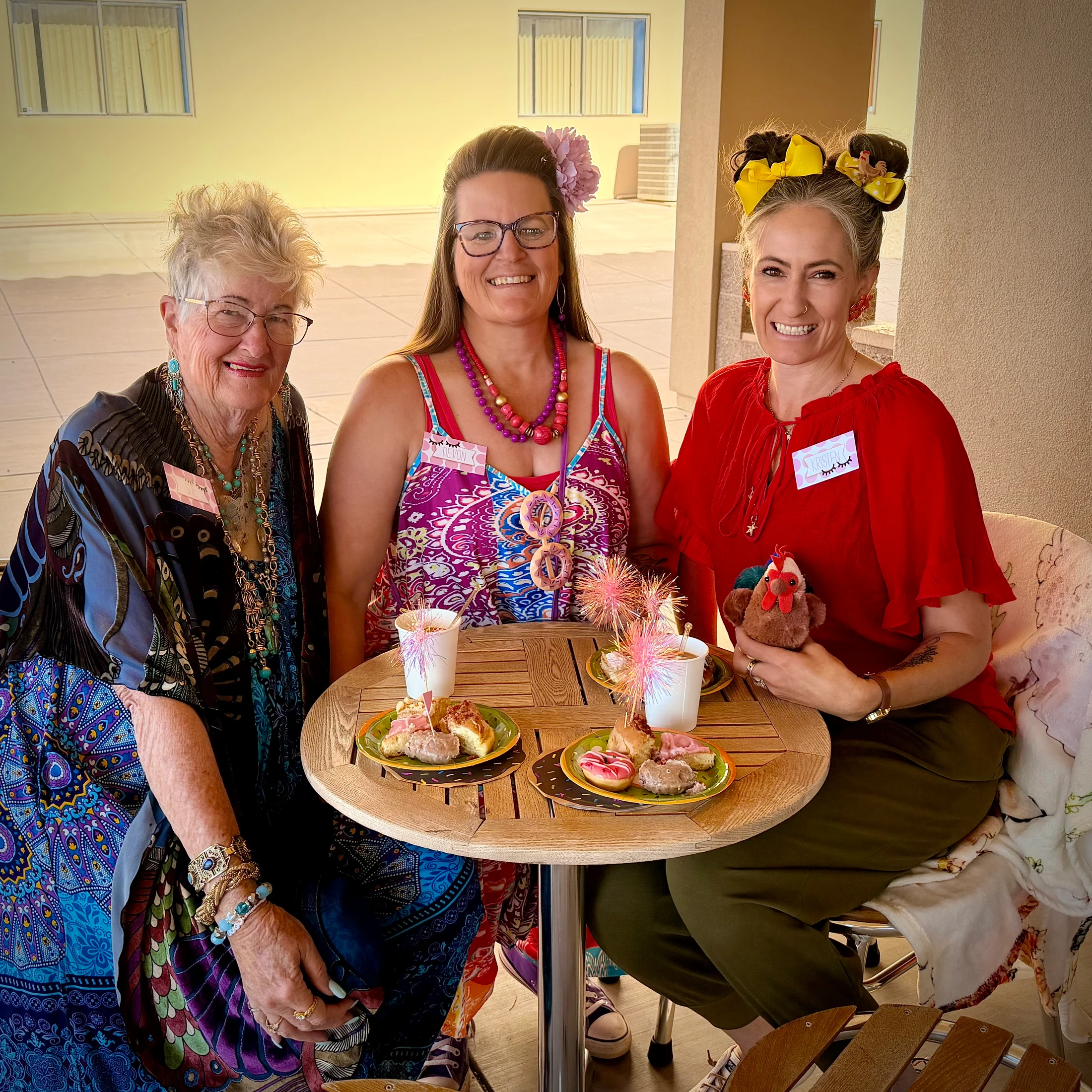 Three smiling women in colorful, festive outfits sit at an outdoor table. On the table are plates of donuts and drinks with sparkly decorations.