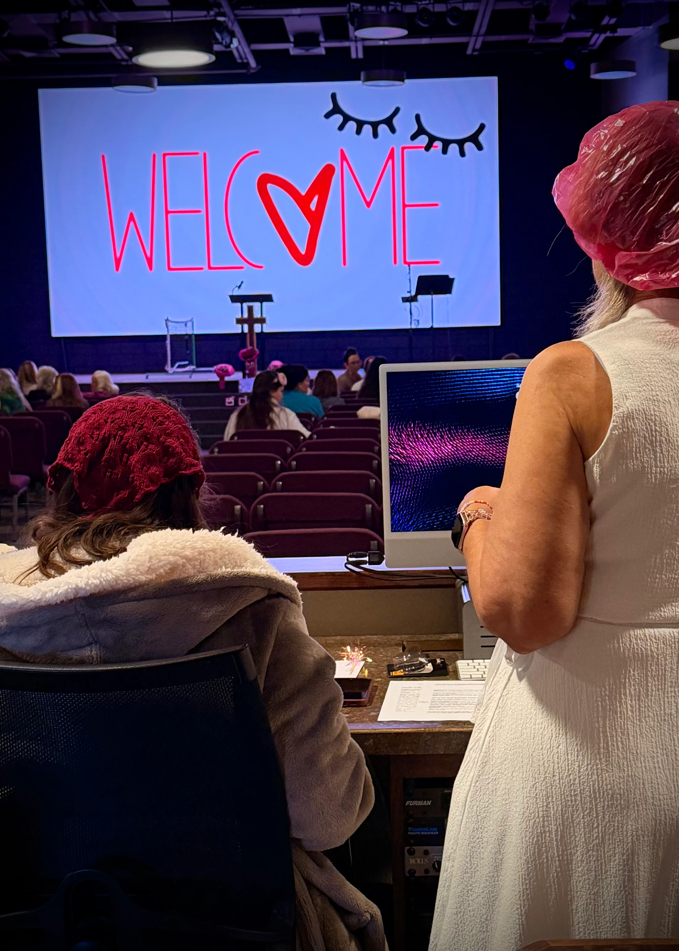 From a tech booth, the view of an auditorium where a large screen on stage reads 'WELCOME' in red text with a heart, facing a seated audience.