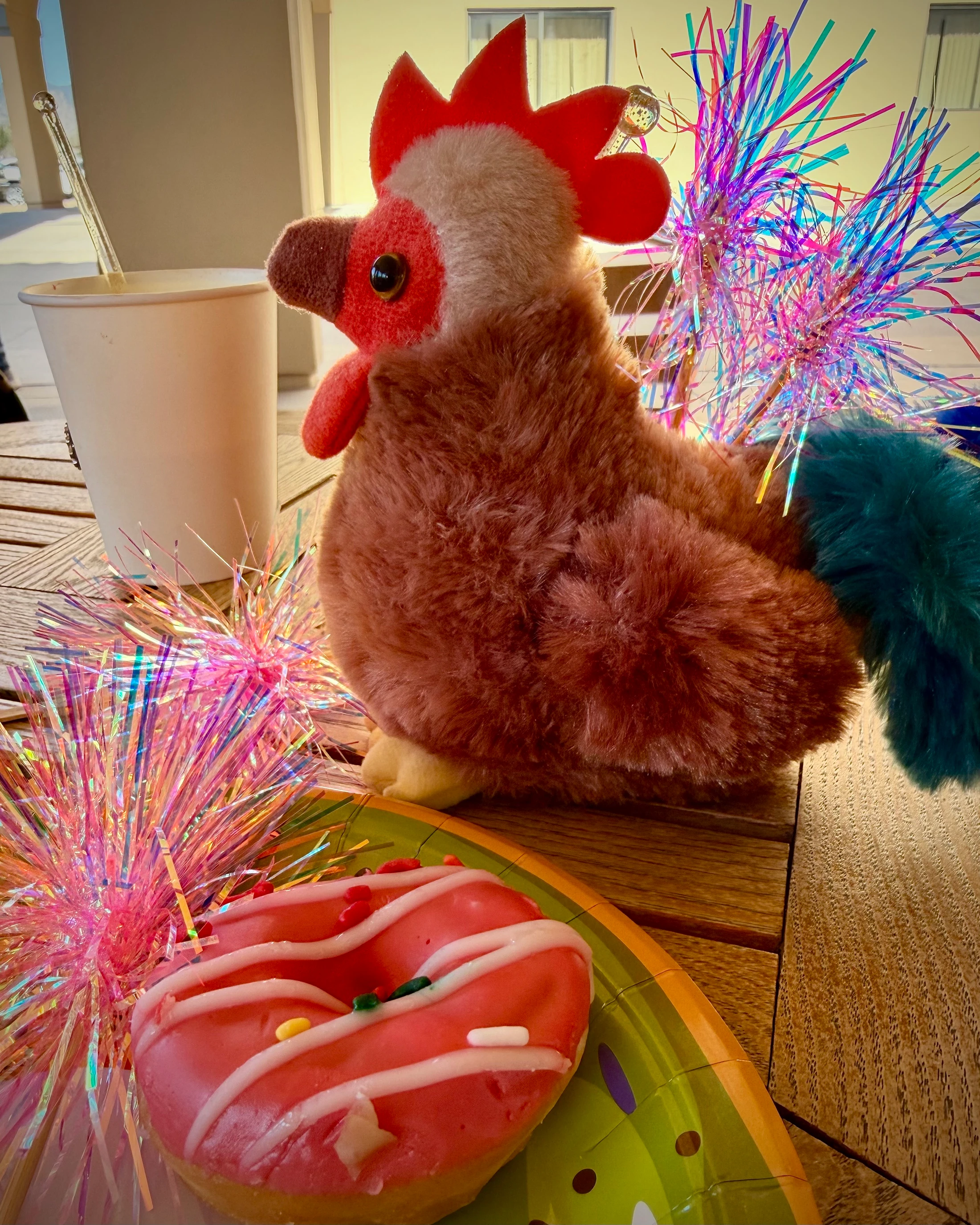 A brown stuffed rooster sits on a wooden table next to a pink donut and a paper cup, surrounded by colorful, iridescent party decorations.