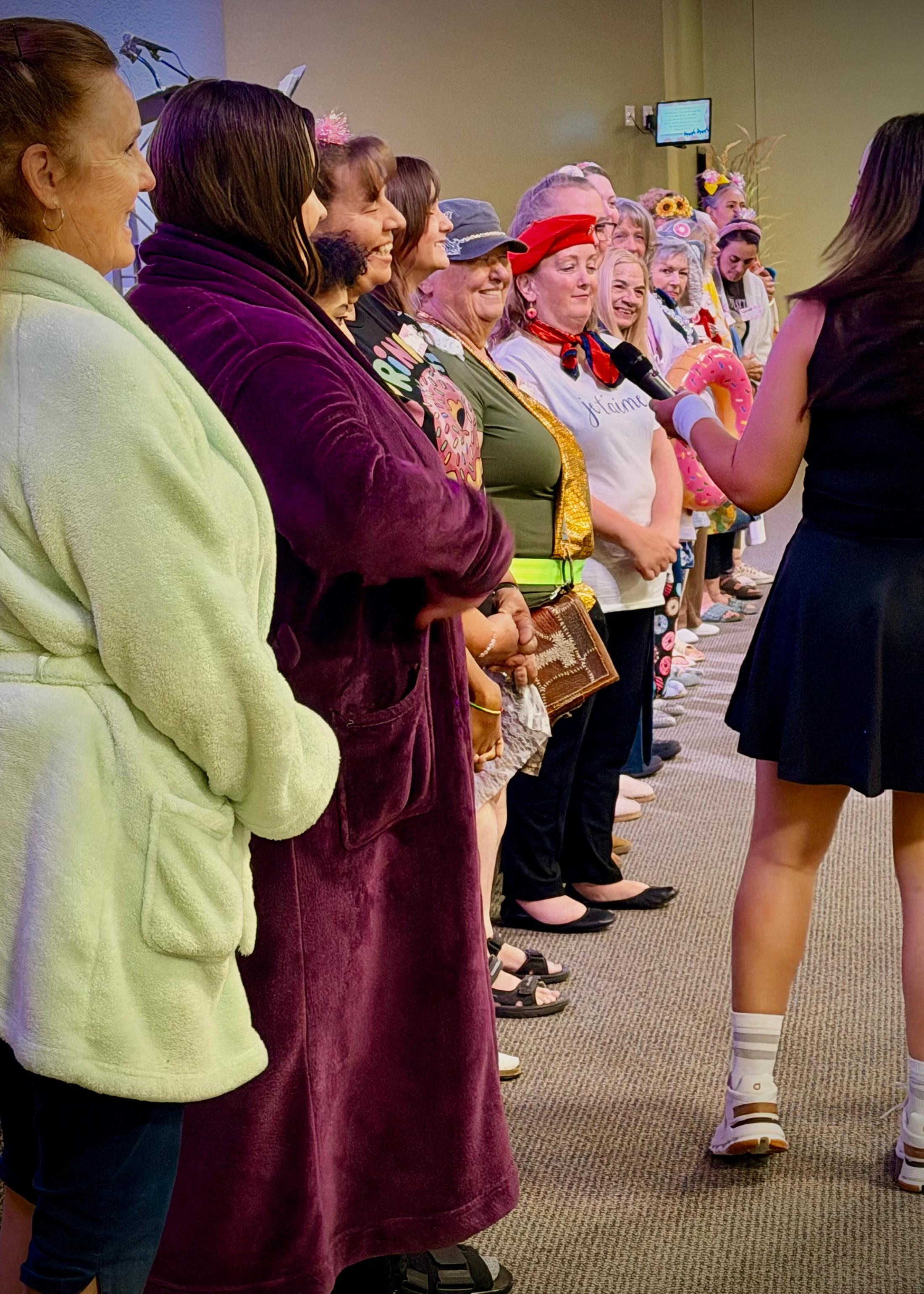 A line of smiling women in fun, eclectic costumes, including bathrobes, stand indoors while a speaker with a microphone addresses them during an event.