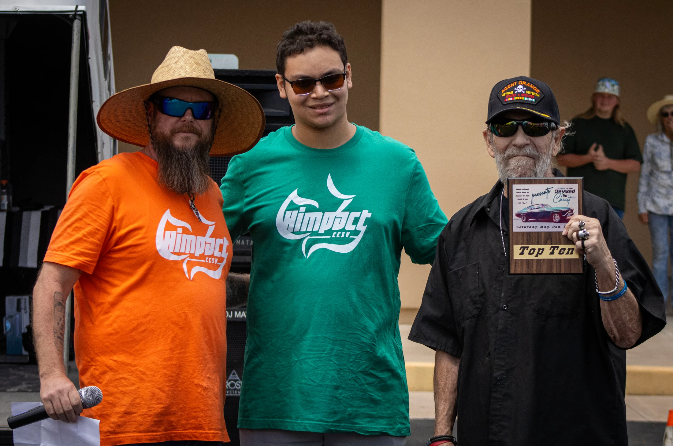 Three men stand together at an event. The man on the right, a veteran, holds a 'Top Ten' award plaque from a car show, flanked by two younger men.