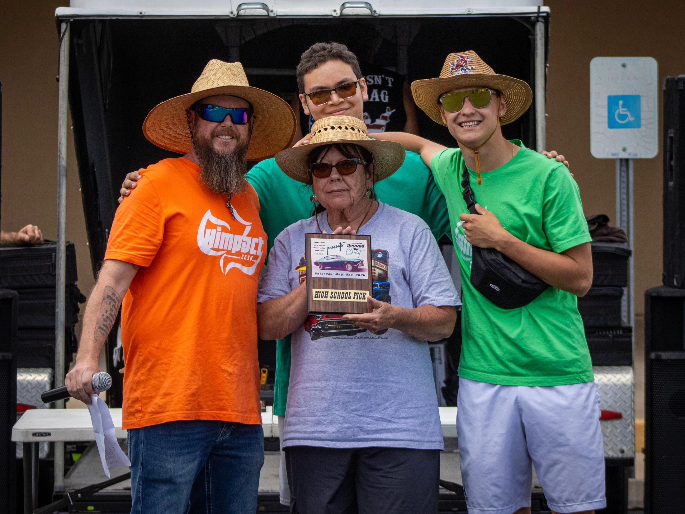 A group of four people, three wearing straw hats, smile for a photo. A woman in the center holds a plaque that reads 'High School Pick'.