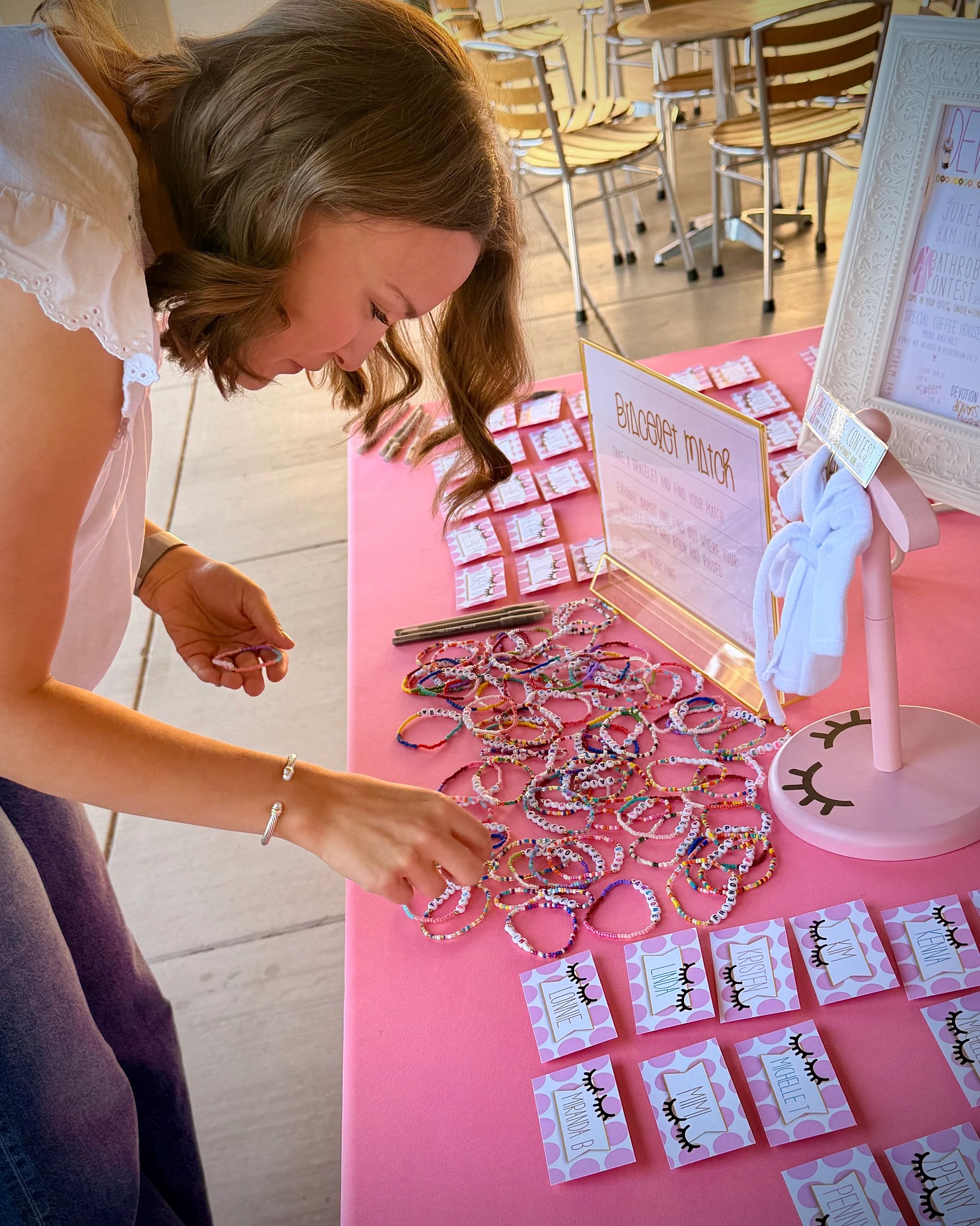 A young woman leans over a pink table, sorting through a large pile of colorful, handmade beaded bracelets with letter beads.