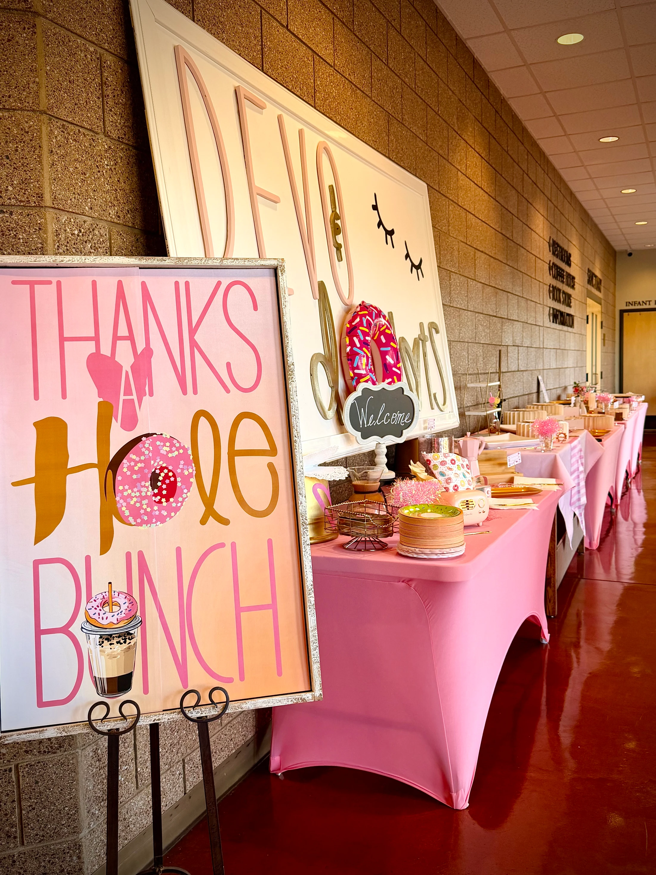 A party setup in a hallway with long tables covered in pink cloths. A sign in the foreground reads 'Thanks a Hole Bunch' with donut and boba tea graphics.