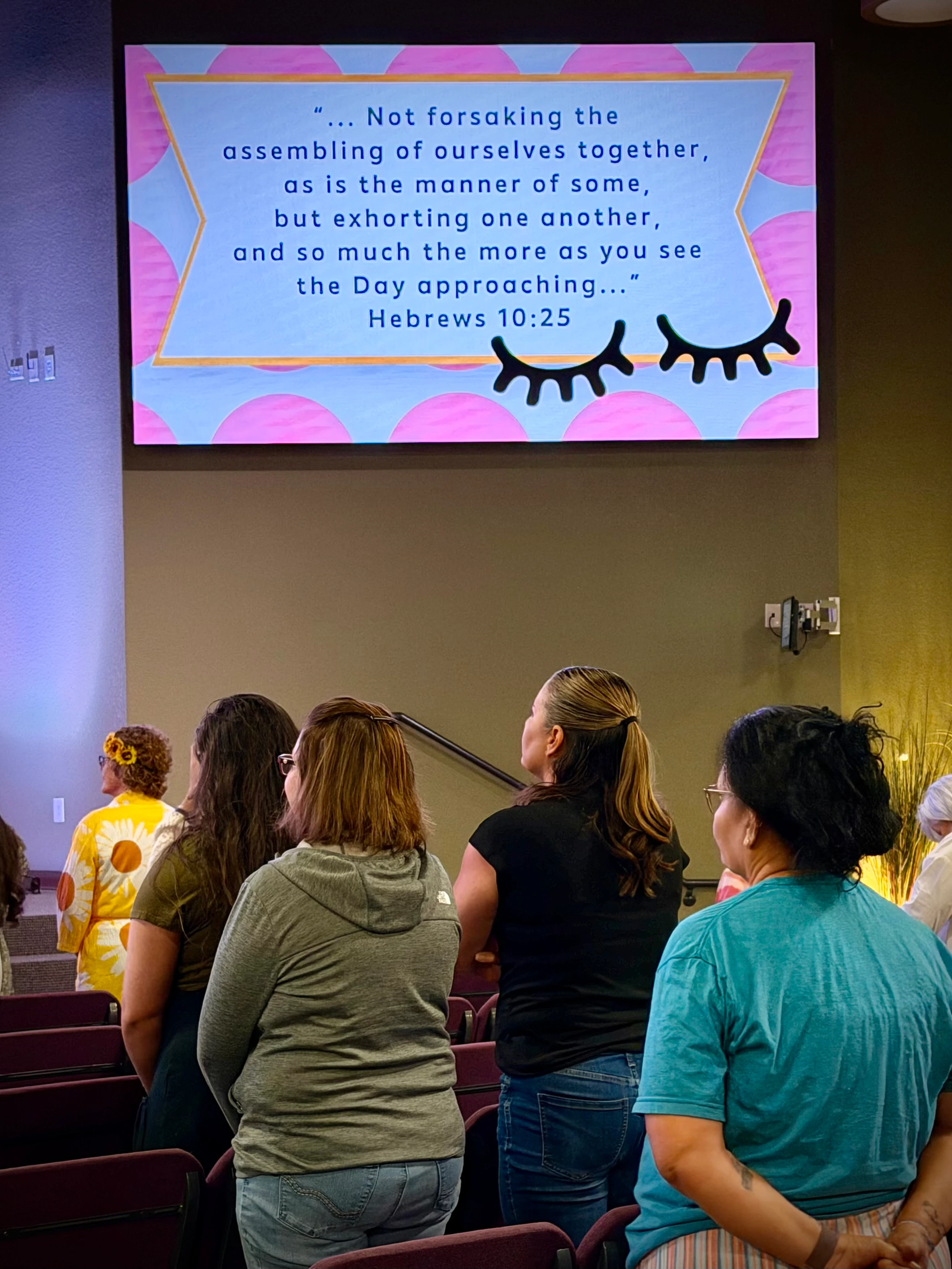 A group of women seen from behind, standing and looking up at a large screen displaying the Bible verse Hebrews 10:25 during a church event.