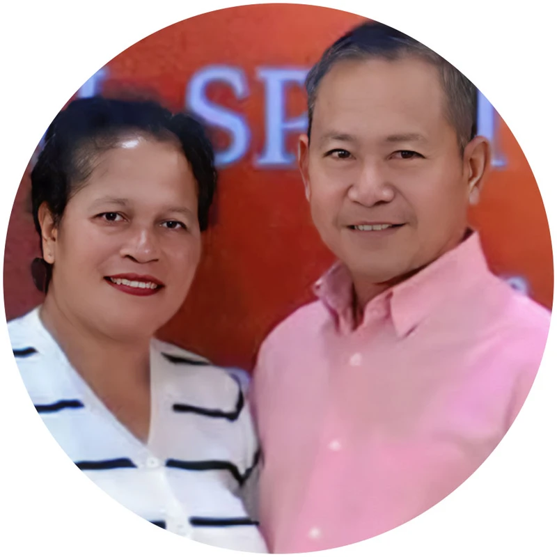 A circular portrait of a smiling middle-aged couple. The woman wears a striped shirt and the man wears a pink button-down shirt against an orange background.