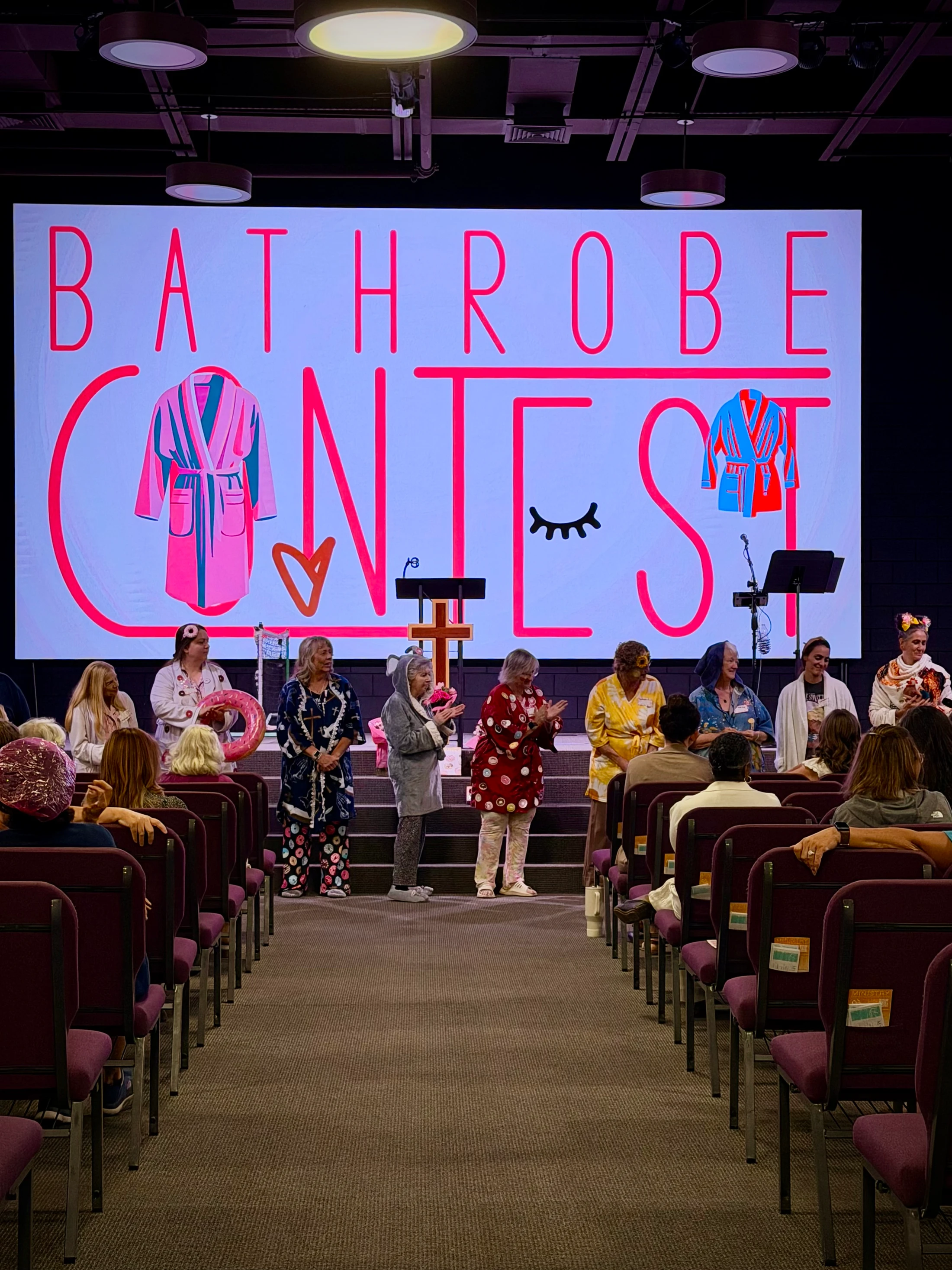 Women in colorful, creative bathrobes stand on a stage for a contest, in front of a large screen that says 'BATHROBE CONTEST', as an audience watches.