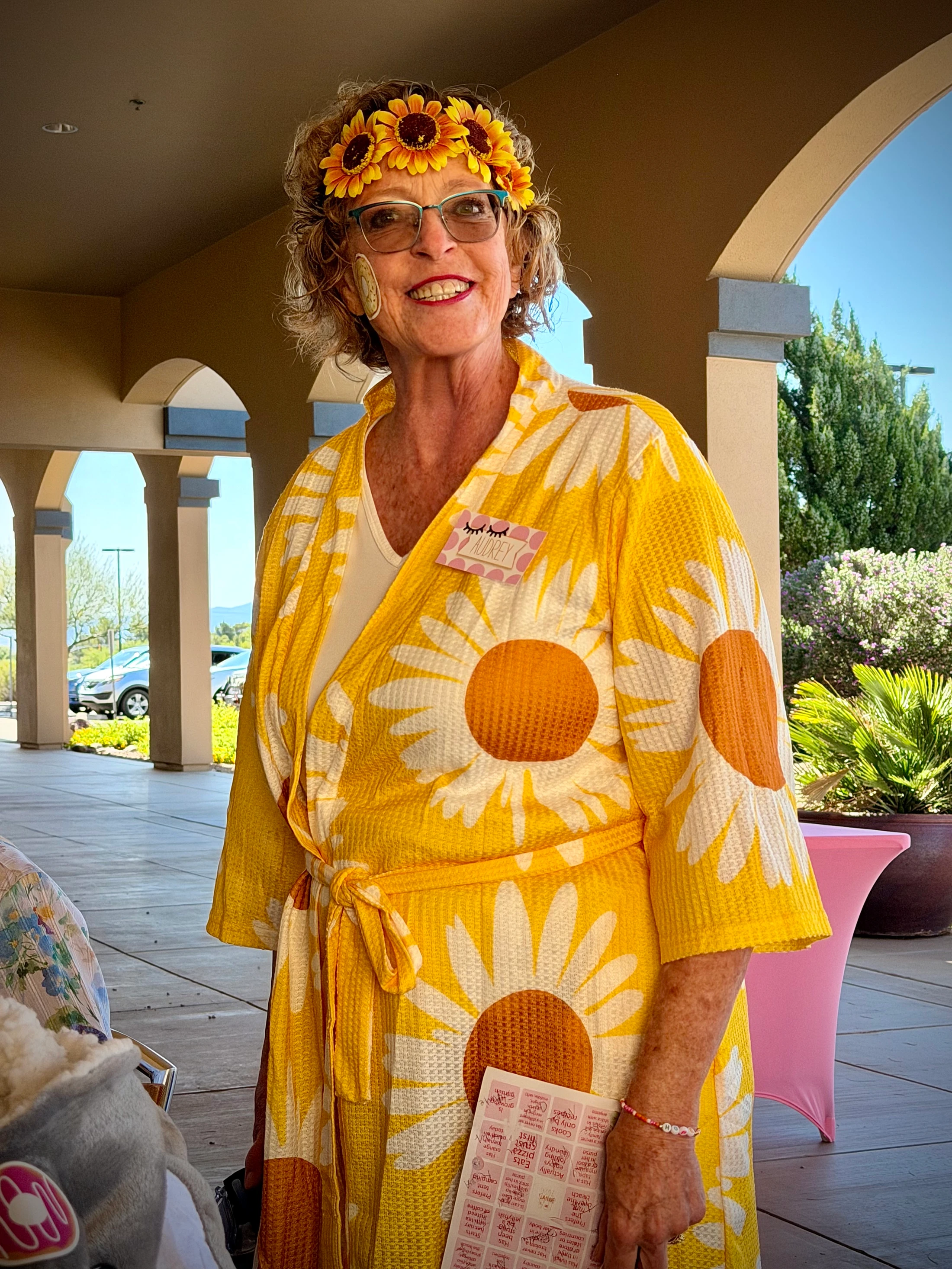 A smiling older woman with glasses wears a yellow daisy-print robe and a sunflower crown. She stands on a sunny patio, looking cheerful at an event.