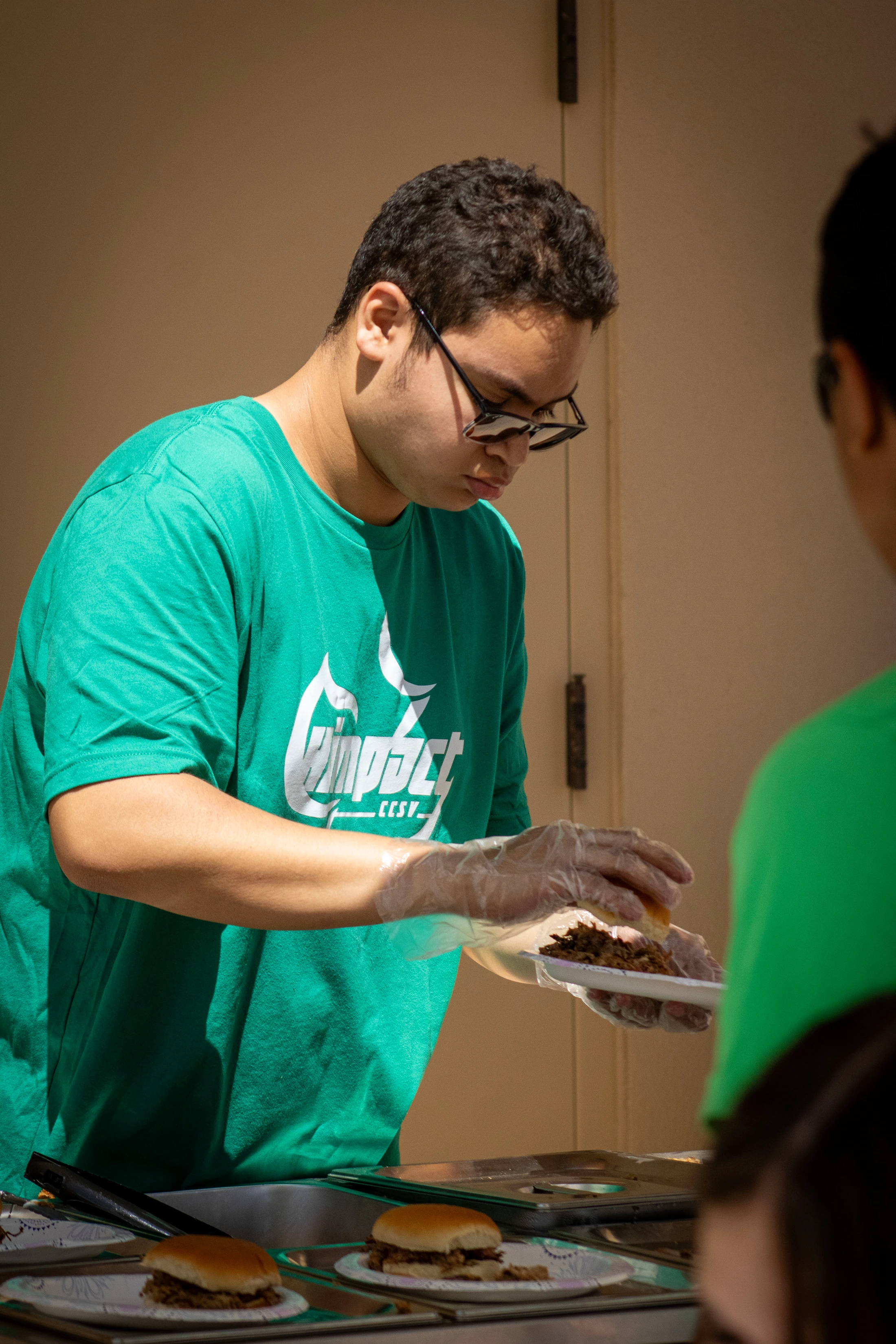 A young man with glasses and a green t-shirt wears gloves while carefully placing a top bun on a small sandwich on a paper plate.