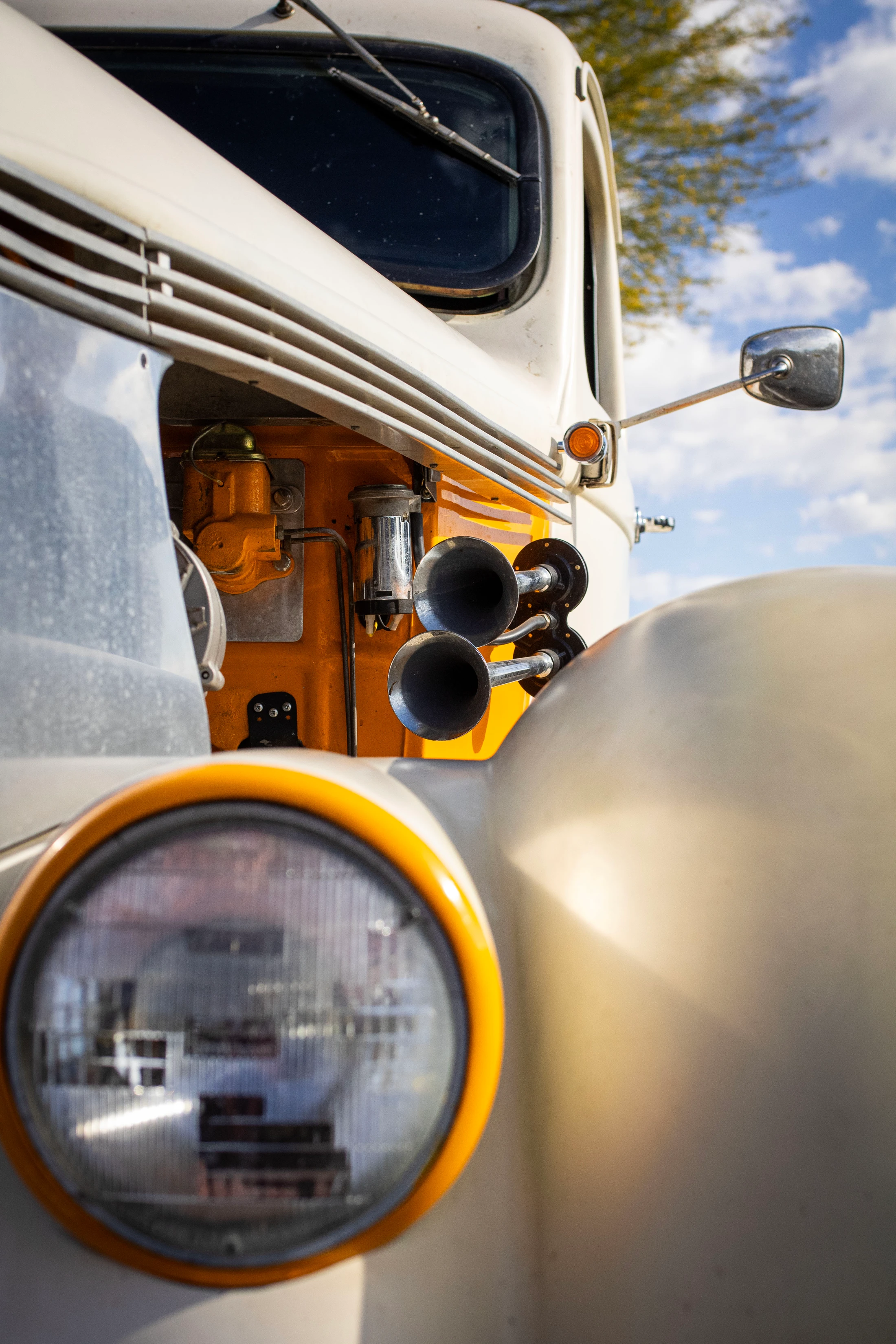 A low-angle close-up of a vintage off-white truck. The headlight, chrome horn, and orange engine block are visible under a blue sky.