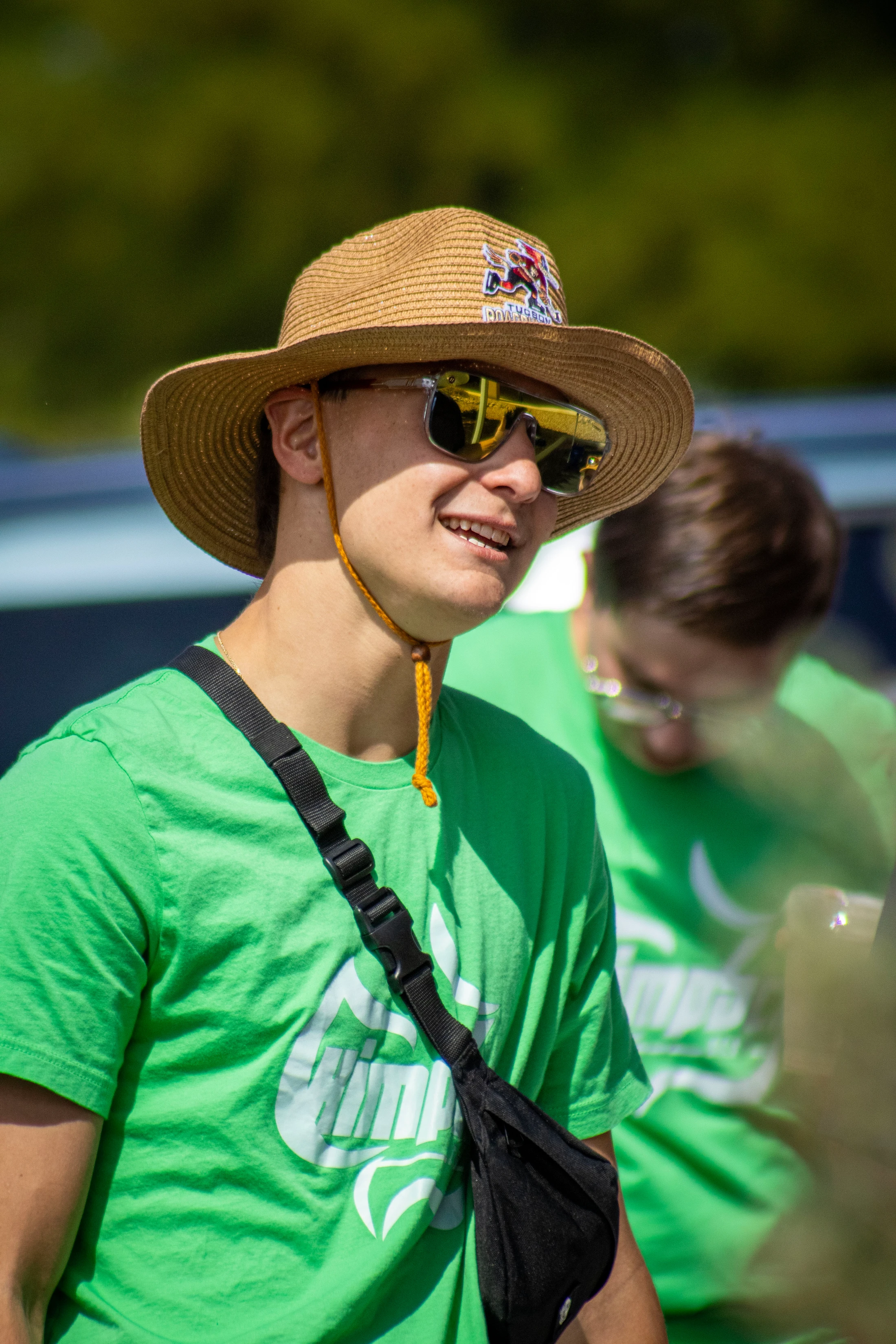 A young man in a green shirt, sunglasses, and a straw hat with a chin strap smiles while looking to his right on a sunny day.