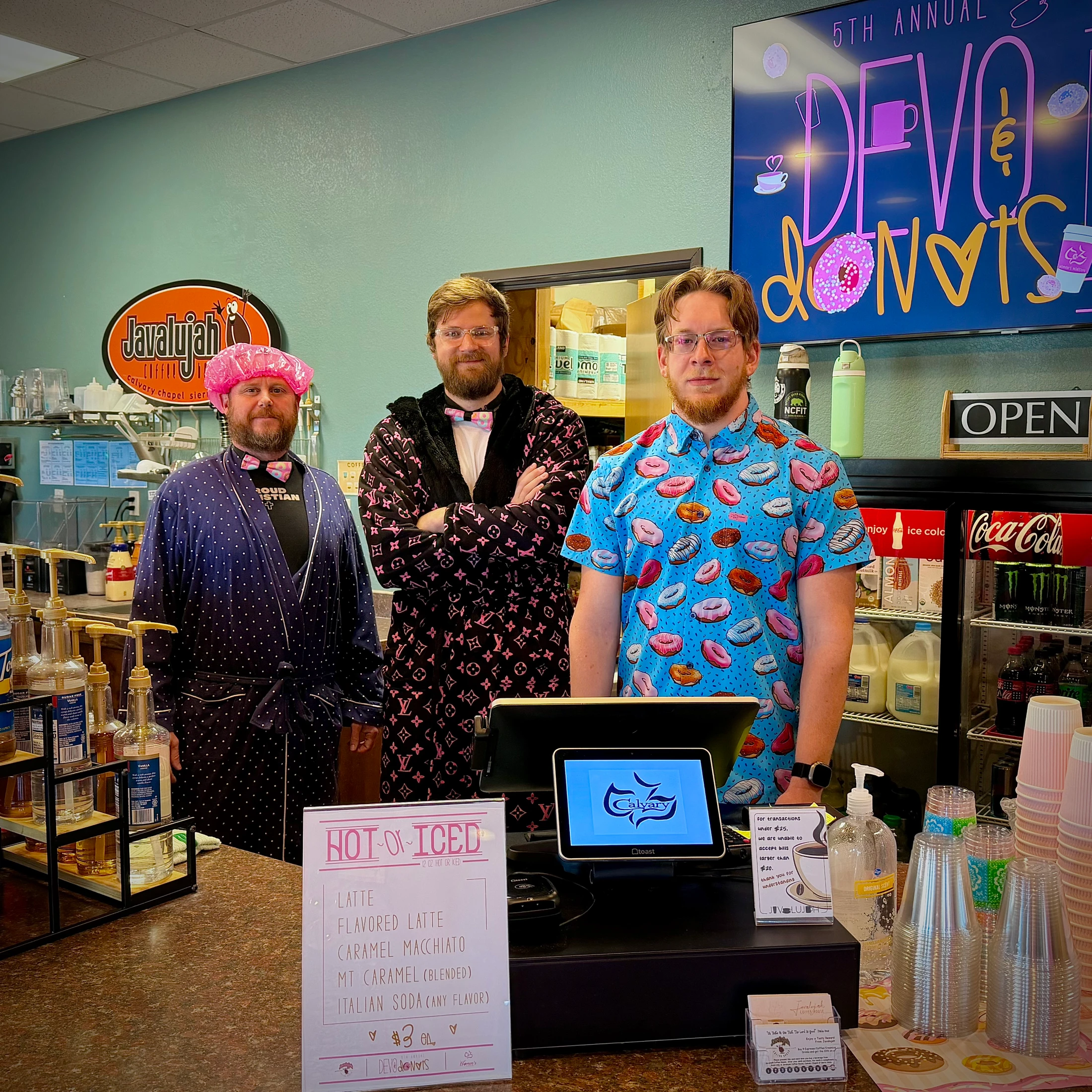 Three men pose behind a coffee shop counter. Two wear bathrobes and bow ties, and the third wears a shirt with a donut pattern for a special event.