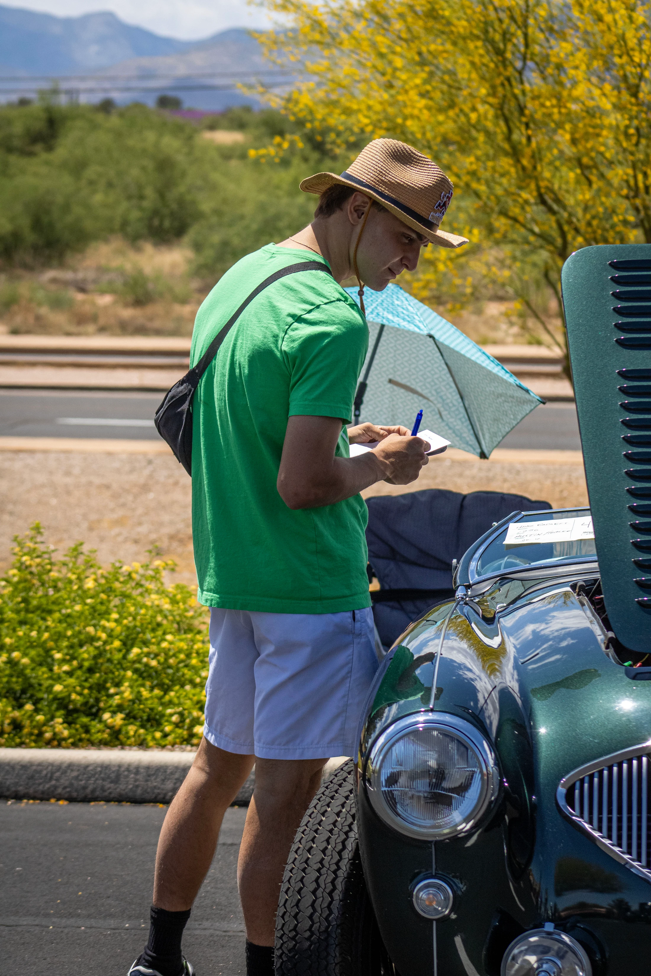 A man in a straw hat and green shirt writes in a notepad while inspecting a vintage dark green convertible car with the hood open at an outdoor event.
