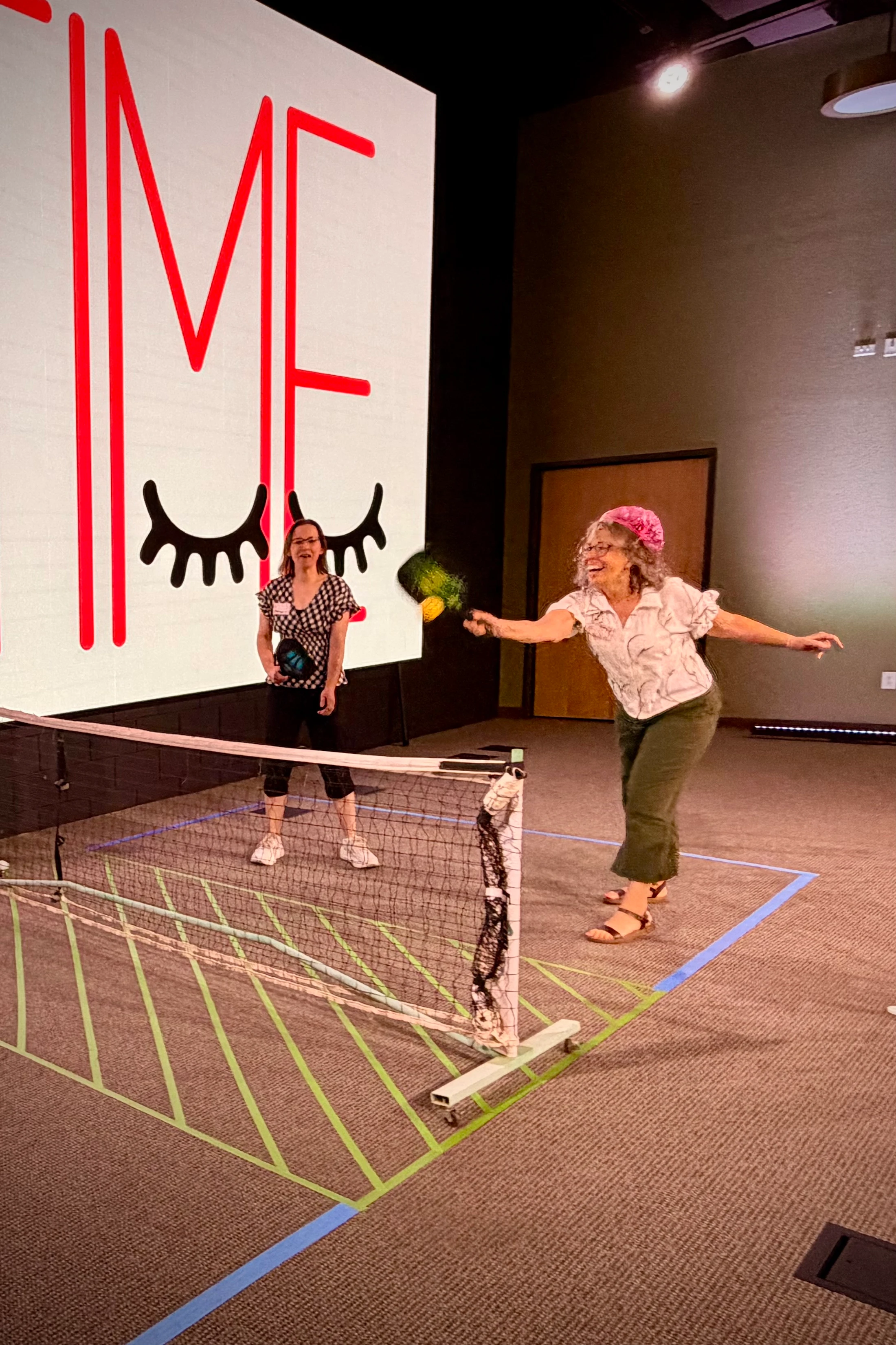 Two women play a game of pickleball on a temporary court set up indoors on a carpet. One woman joyfully hits the ball as the other watches from across the net.
