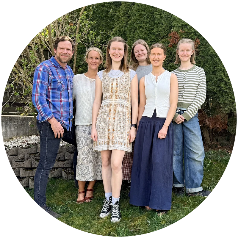 A family of six, including parents and four daughters, stands together smiling for a photo in a grassy backyard with green bushes behind them.