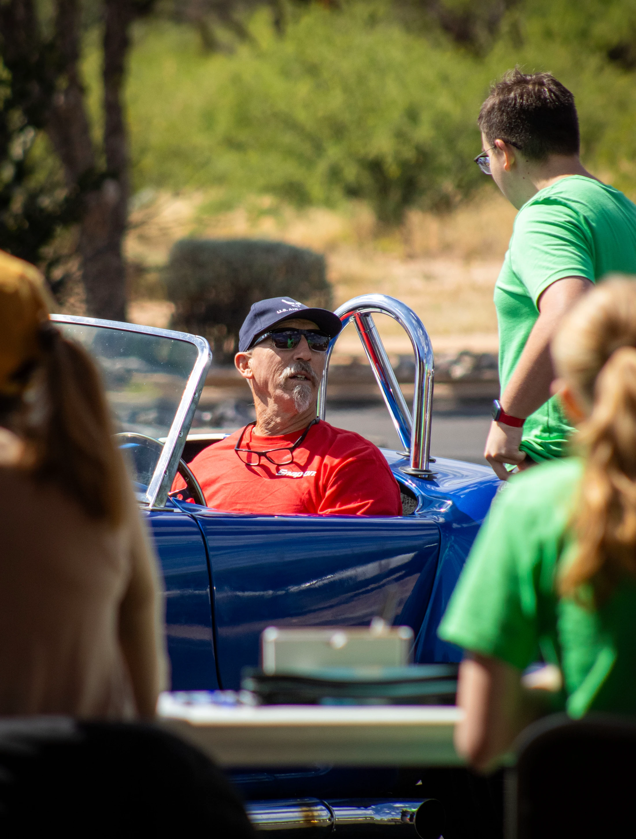 An older man with a goatee and sunglasses sits in the driver's seat of a blue convertible, talking to a younger man standing next to the car outdoors.