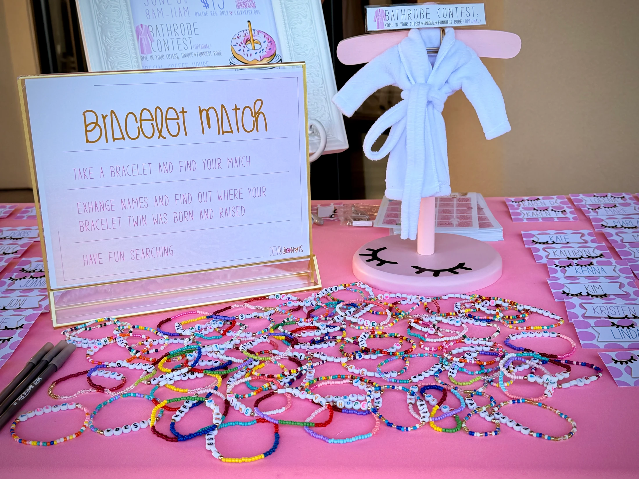 A pink table is set up for a party game called 'Bracelet Match' with a large pile of colorful, handmade beaded bracelets with words on them.