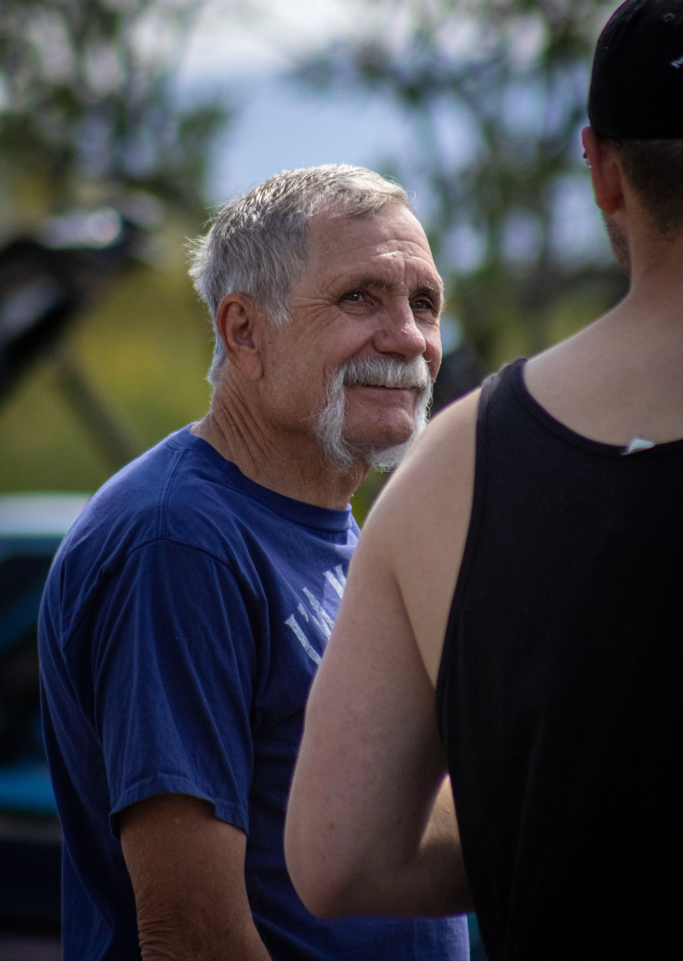 An older white man with grey hair and a large handlebar mustache smiles while talking to another person. He wears a blue t-shirt and is seen from the chest up.