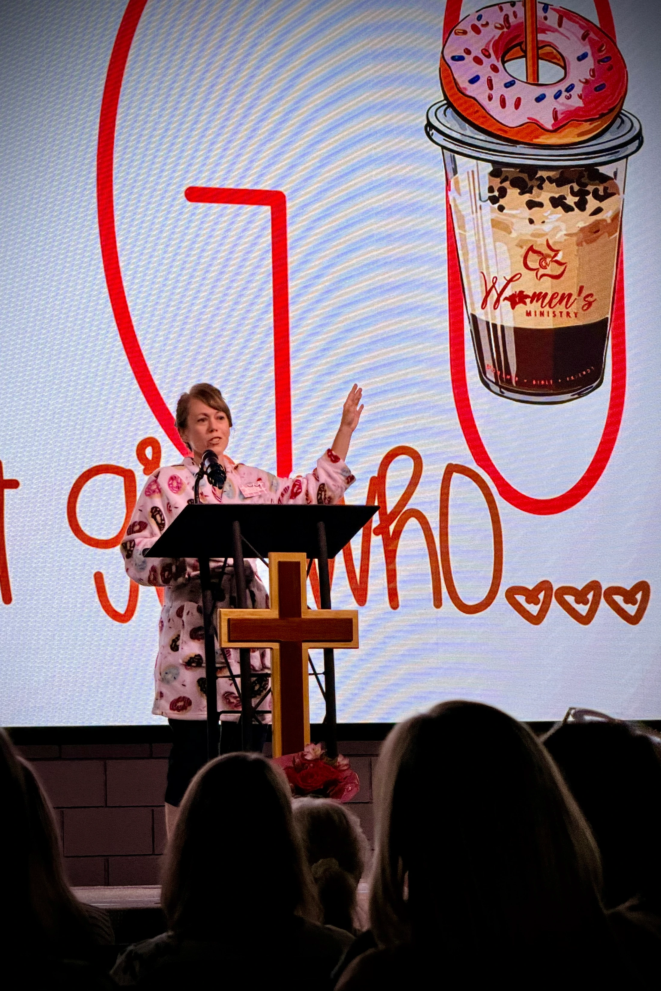A woman in a donut-print robe speaks from a podium with a cross. Behind her, a screen displays a graphic for a women's ministry event with coffee and a donut.