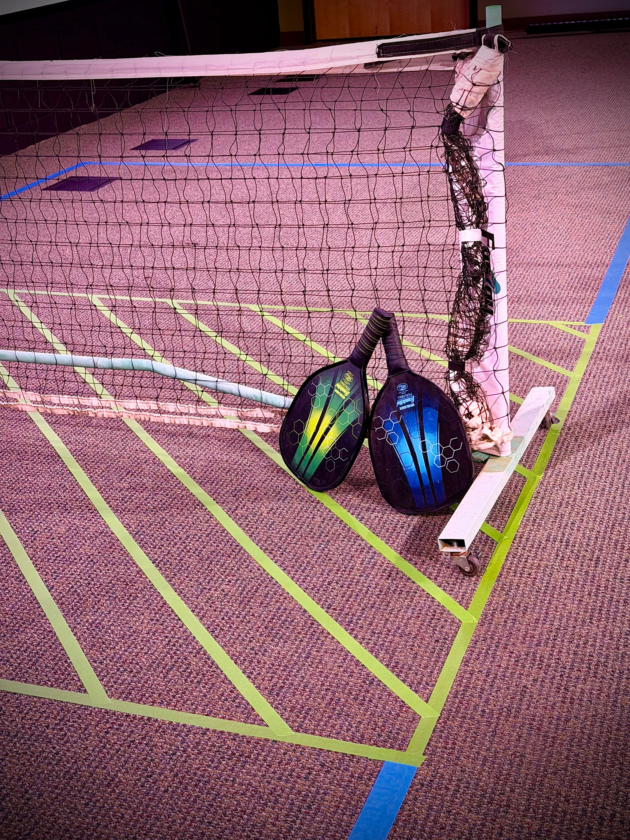 Two pickleball paddles, one green and one blue, leaning against a net on a carpeted floor. The court lines are marked out with green and blue tape.
