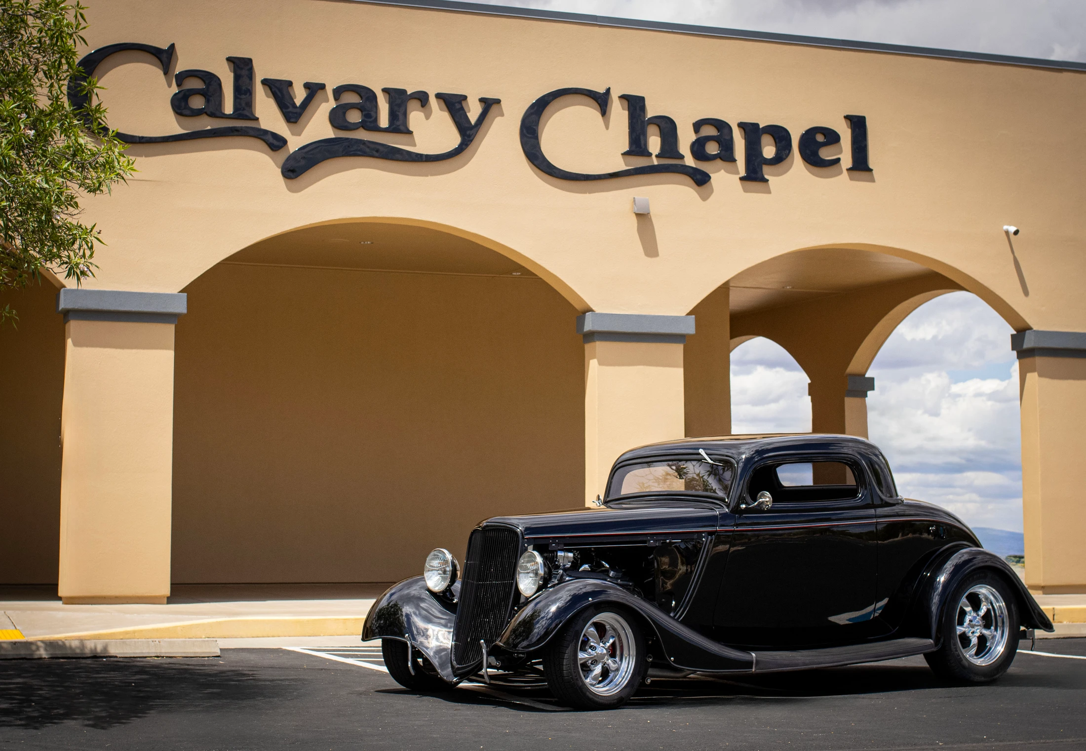 A shiny, classic black hot rod coupe is parked in the foreground of a tan building with a sign that reads "Calvary Chapel".