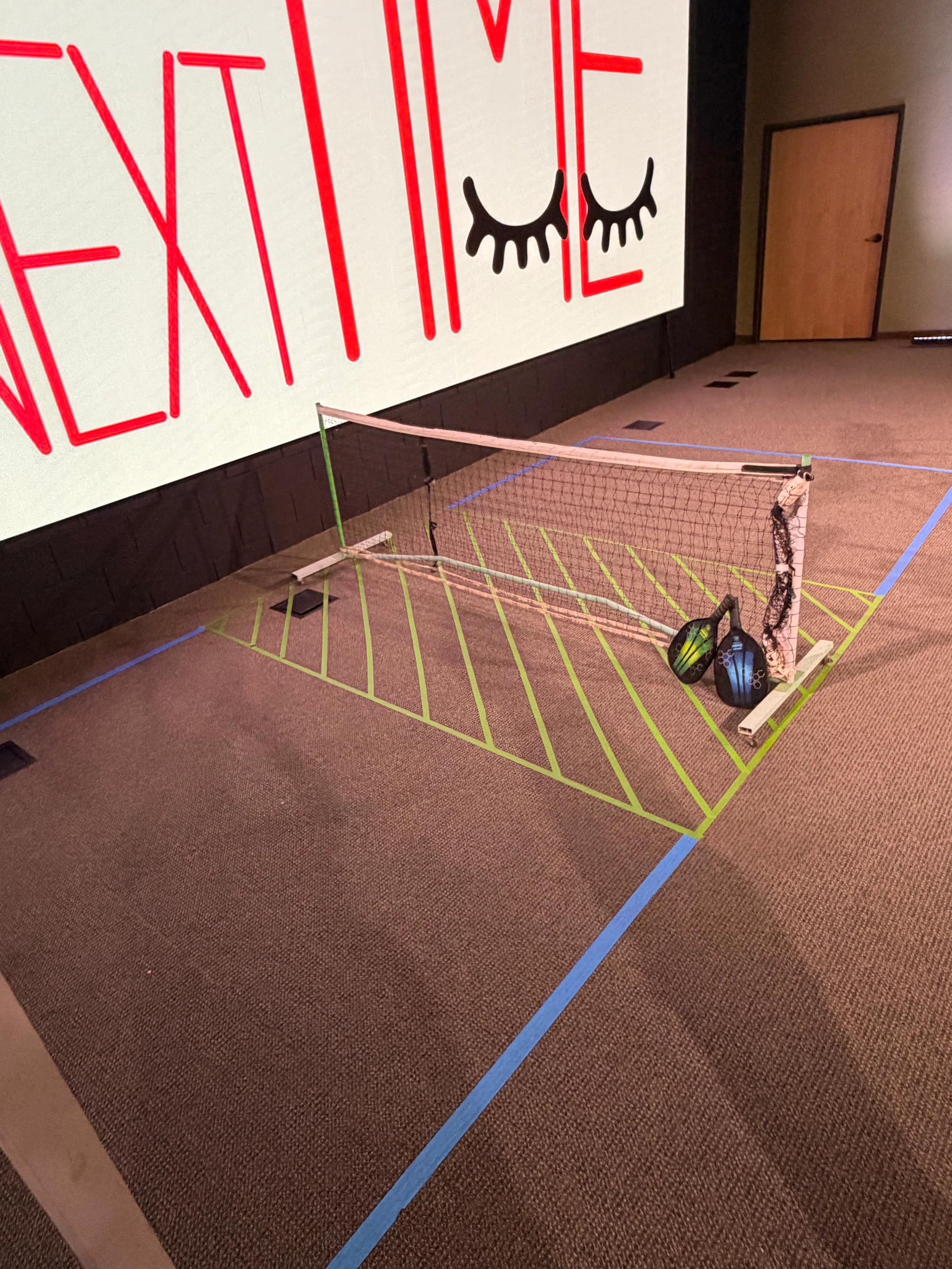 A small pickleball court is set up indoors on a brown carpet using tape for lines. Two paddles rest against the net, in front of a large screen.