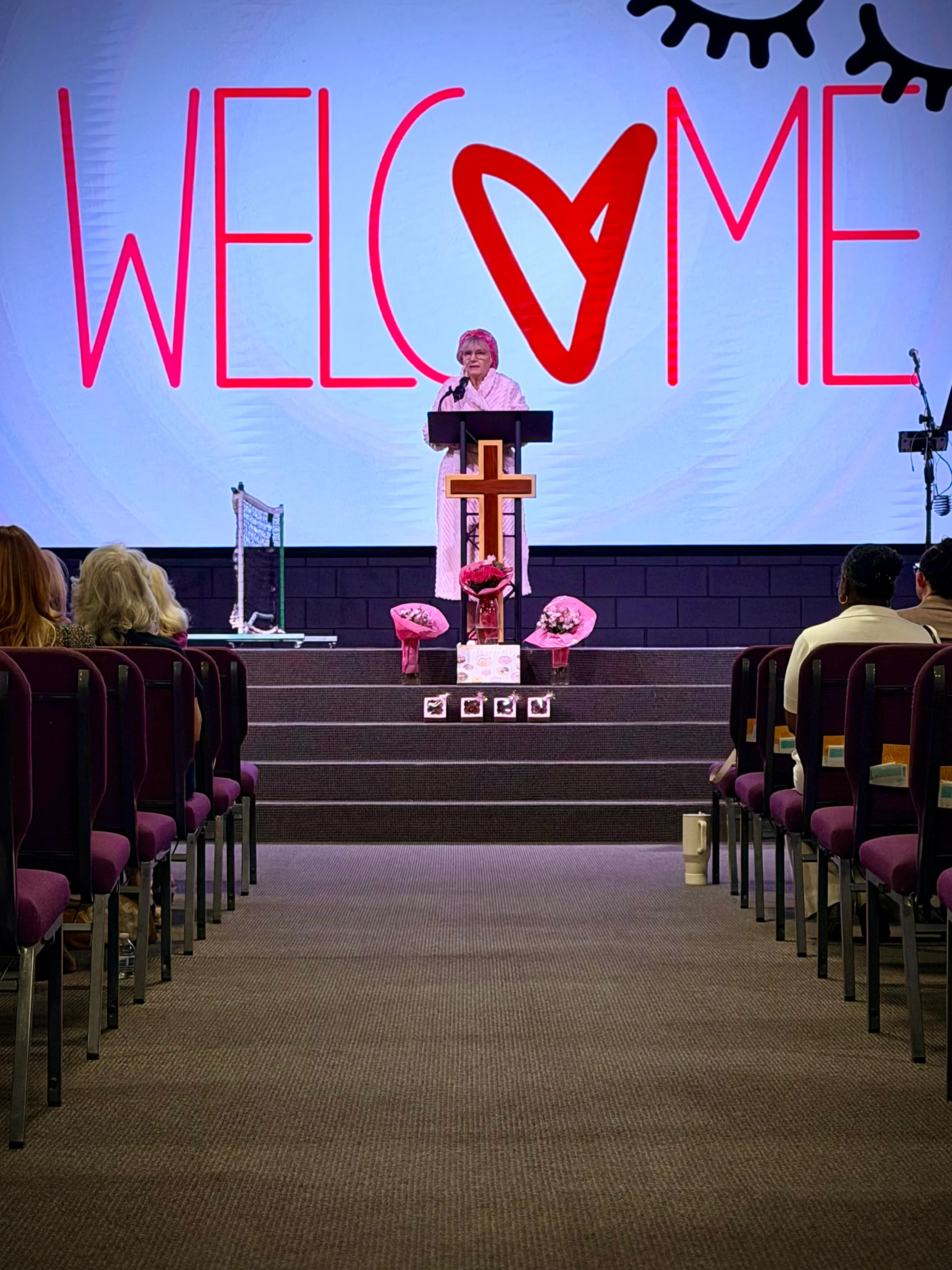 An older woman speaks at a podium on a church stage. Behind her, a large screen displays the word 'WELCOME' in red with a heart for the letter O.