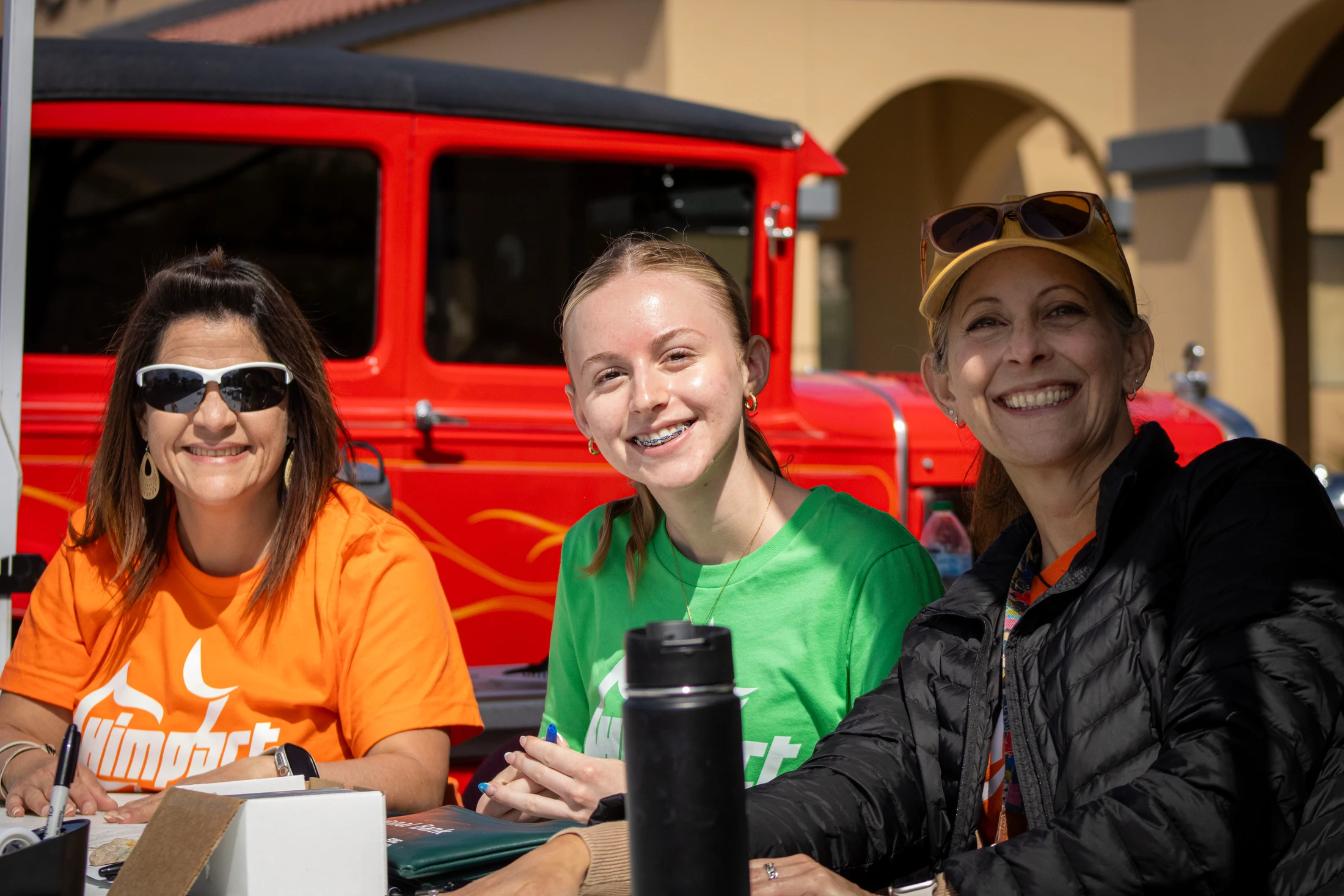 Three women sit at a table outdoors, smiling for the camera. They appear to be volunteers at an event, with a large red vintage truck behind them.
