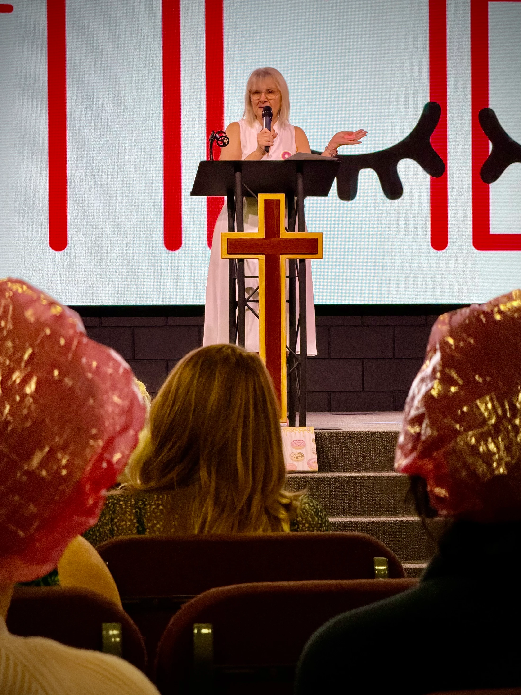 A woman with glasses speaks at a podium adorned with a large cross. In the foreground, audience members wear shiny pink hats.