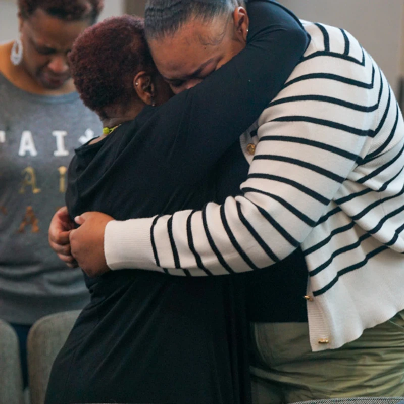 Two Black women hug tightly in an emotional embrace. In the background, another woman with short hair bows her head in what appears to be a church or service.