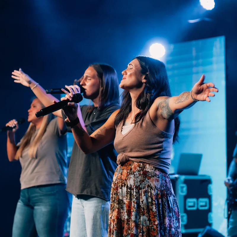 Three women sing into microphones on a stage with blue lights. The lead singer has her arms outstretched, and a guitarist plays in the background.