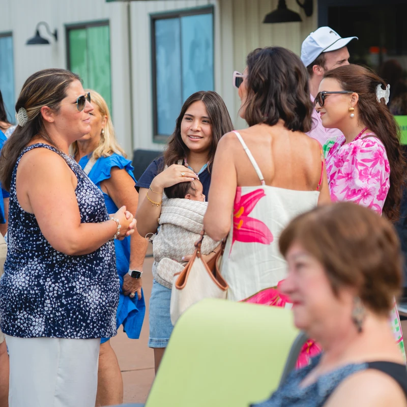 A group of casually dressed people stand and chat outdoors. In the center, a young woman holding a baby in a carrier smiles at the woman next to her.