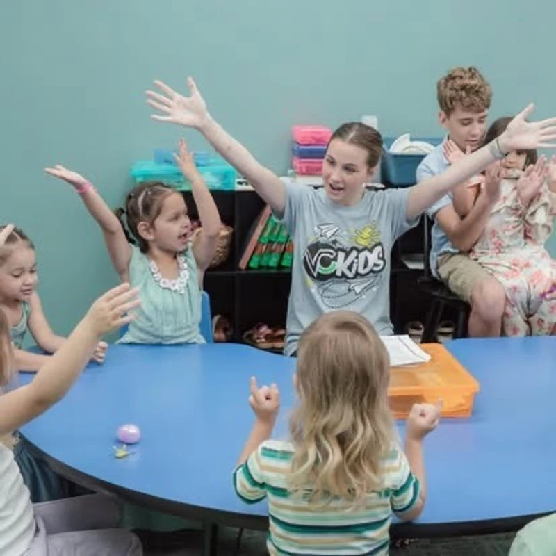 A female teacher leads a group of young children in an activity around a blue table, with many of them raising their arms and singing joyfully.