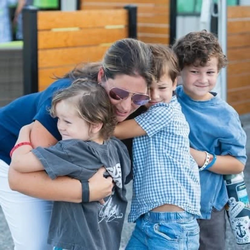 A smiling woman wearing sunglasses shares a warm group hug with her three young sons outdoors.