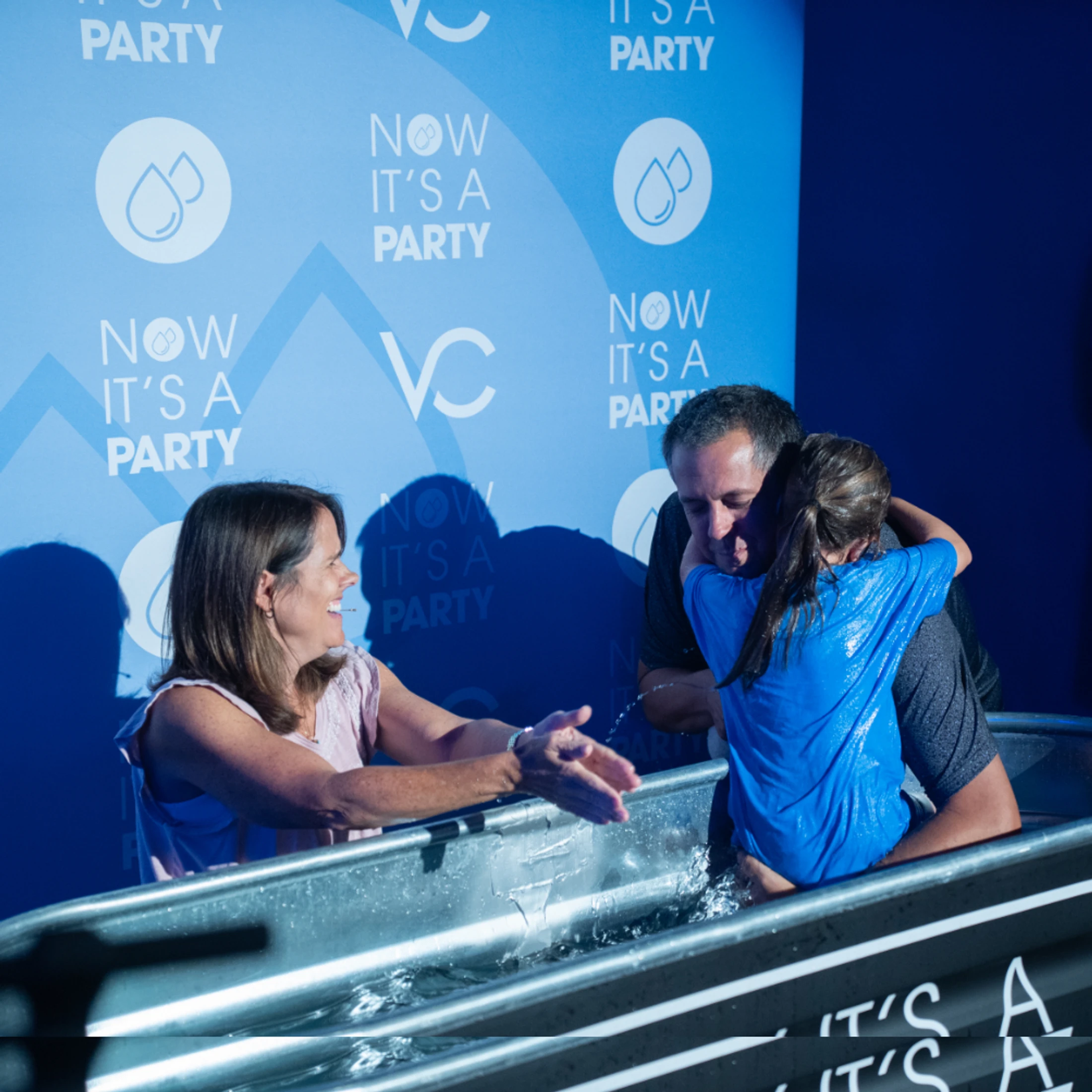 A man hugs a wet young person in a baptismal tub as a woman smiles and reaches toward them. The backdrop is blue and reads, "Now It's A Party."