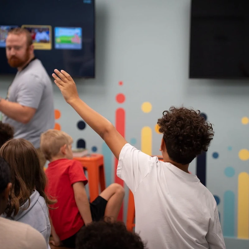 A young student with curly hair, seen from the back, raises their hand to ask a question as a teacher leads a lesson for a group of children in a classroom.