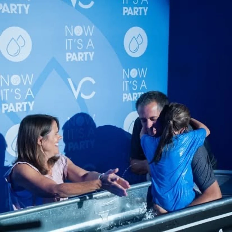 A man holds a young girl in a baptismal pool as she hugs him. A woman smiles at them from the side. A blue banner behind them reads, "Now It's a Party."