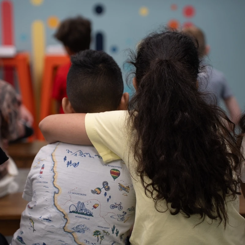 From behind, a girl with long dark hair has her arm around a boy as they sit with other children in a colorful classroom setting, facing away from the camera.