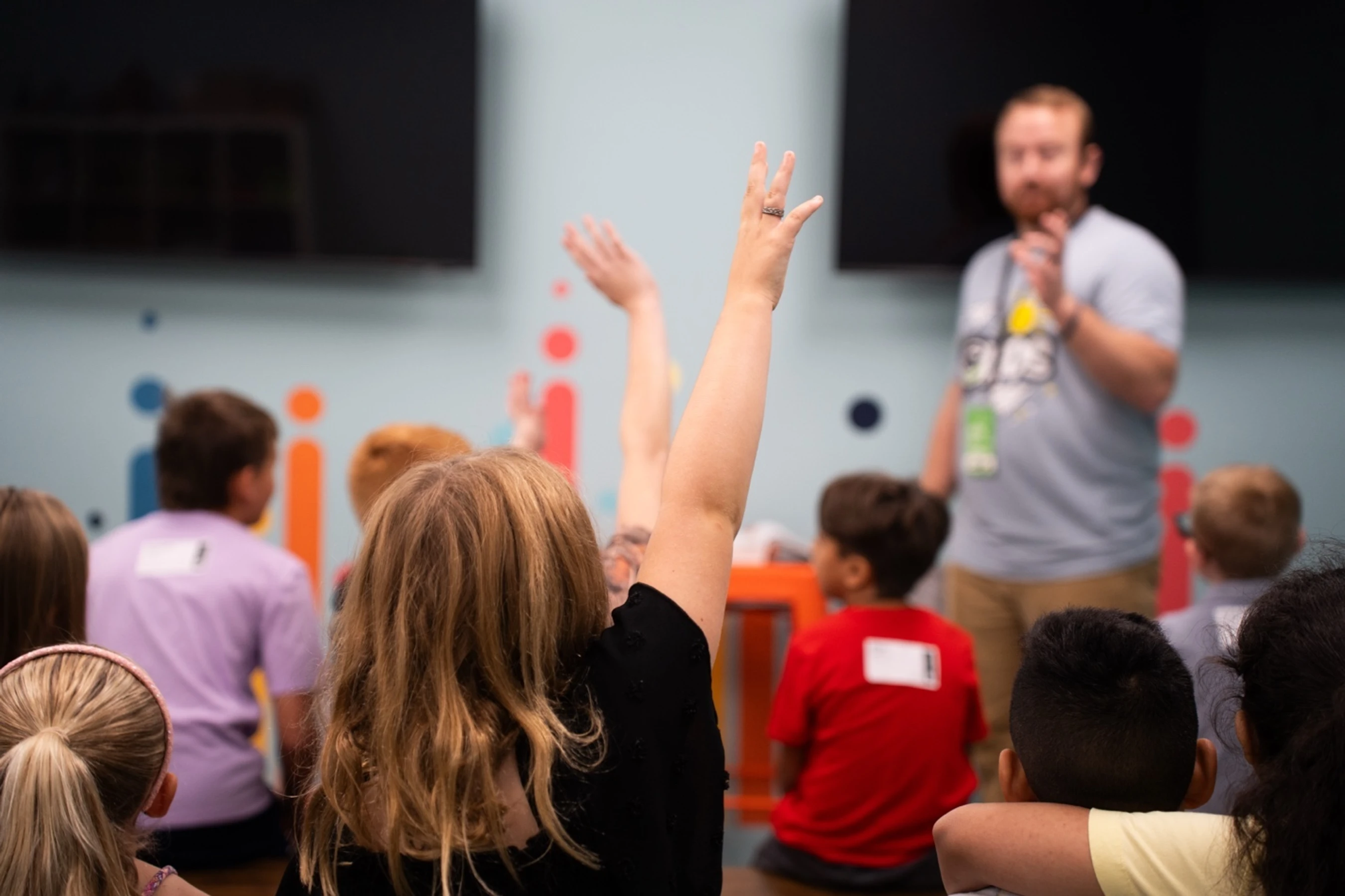 A view from behind of children in a classroom. One person's hand is raised high, with a male teacher out of focus in the background.