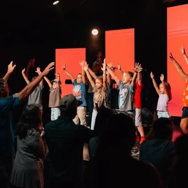A group of children and adults stand on a stage with a red backdrop, raising their hands in worship while an audience watches from the foreground.