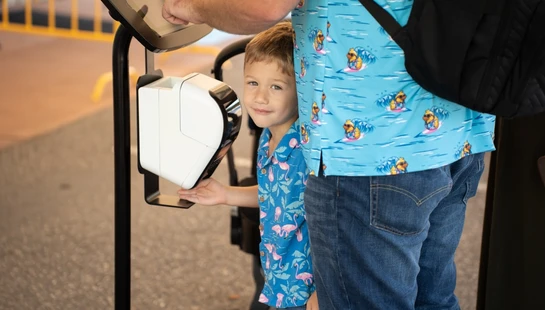 A young boy looks at the camera while his father uses a touchscreen kiosk. The boy has his hand on the scanner below the screen.
