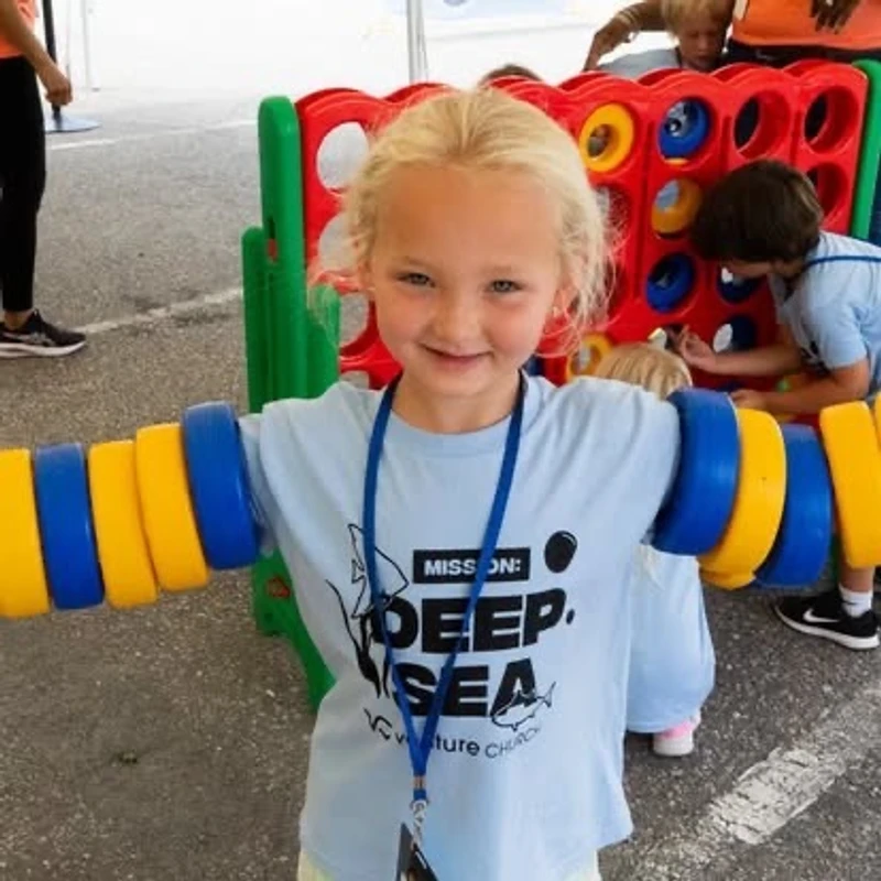 A young blonde girl in a blue 'Deep Sea' t-shirt smiles with large yellow and blue plastic rings on her arms, with a giant Connect Four game behind her.