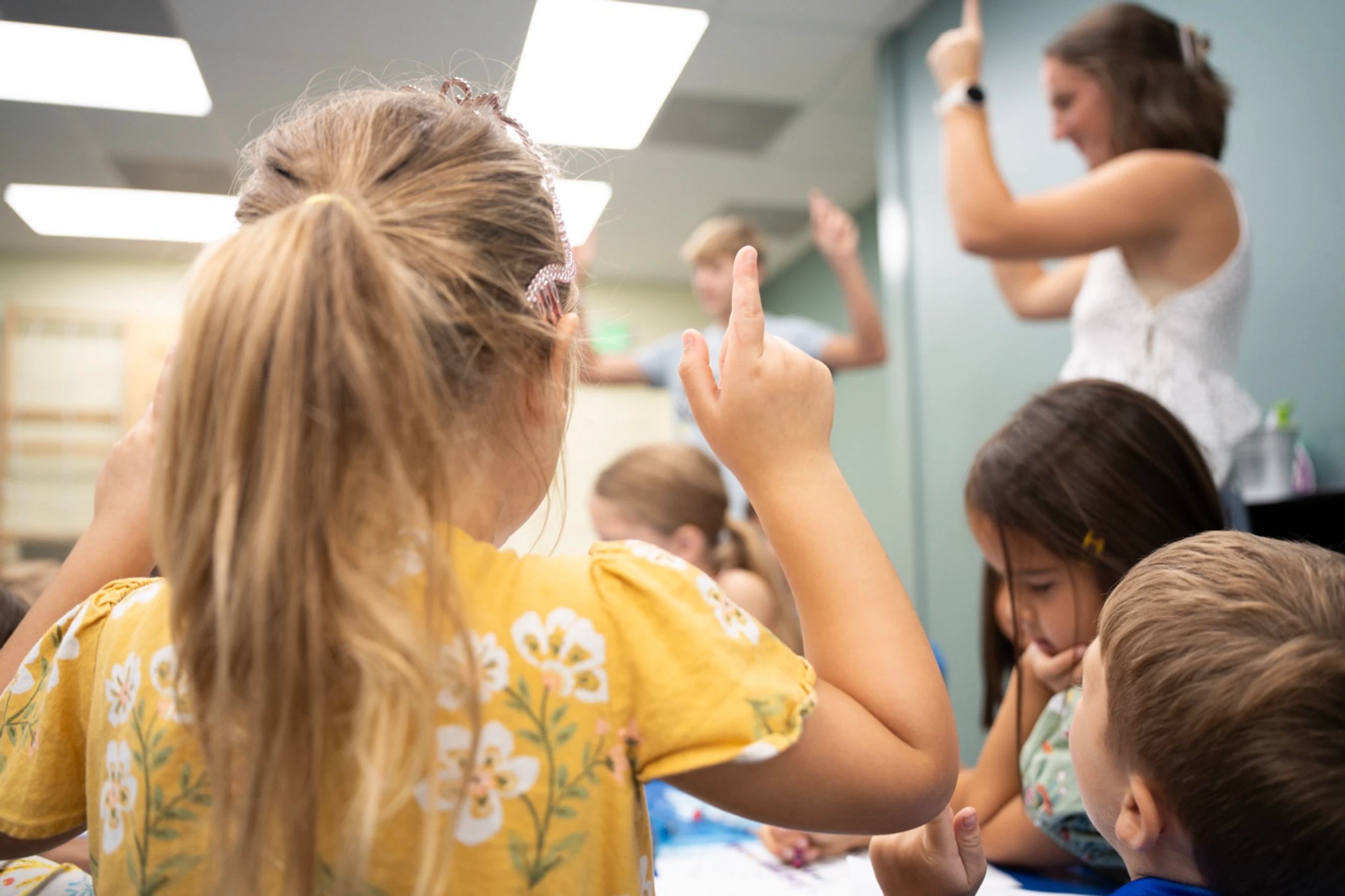 A view from behind a young girl with blonde hair in a yellow floral shirt raising her hand in a classroom, with other students and a teacher in the background.